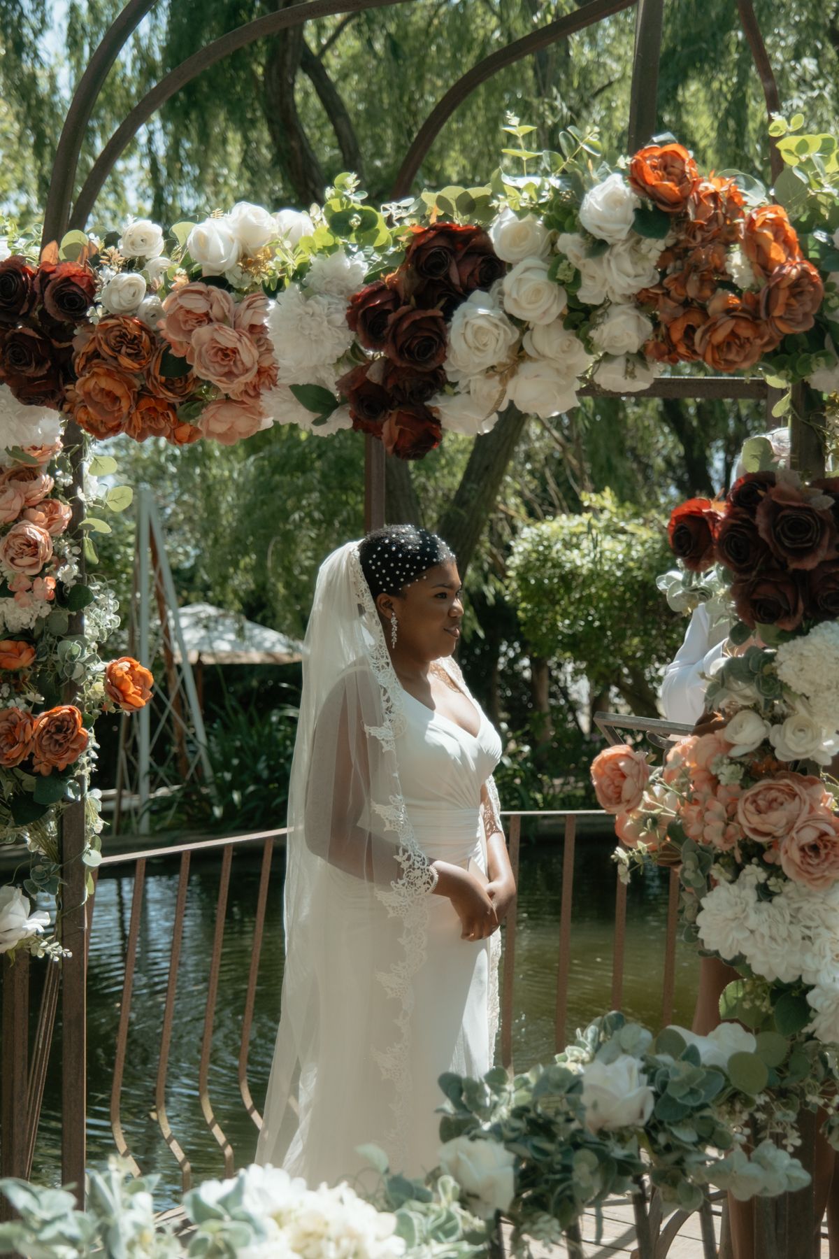 Bride in white gown and veil stands beneath floral arch adorned with orange, red, and white roses.