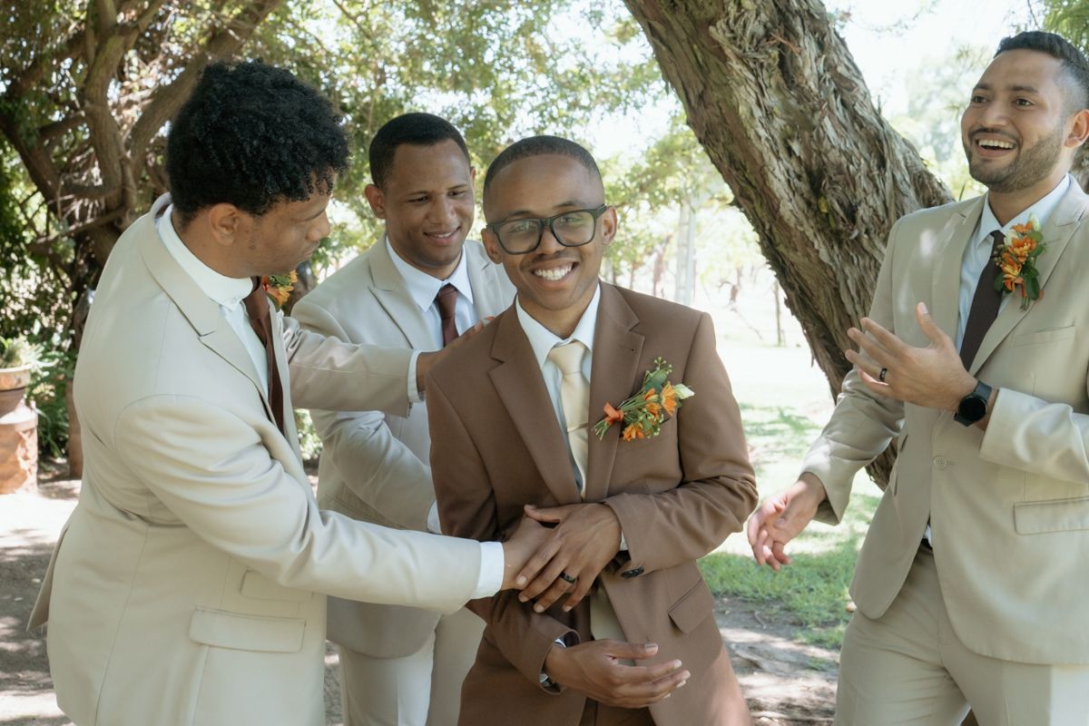 Groom in brown suit laughs with groomsmen in beige suits during a joyful outdoor wedding celebration.