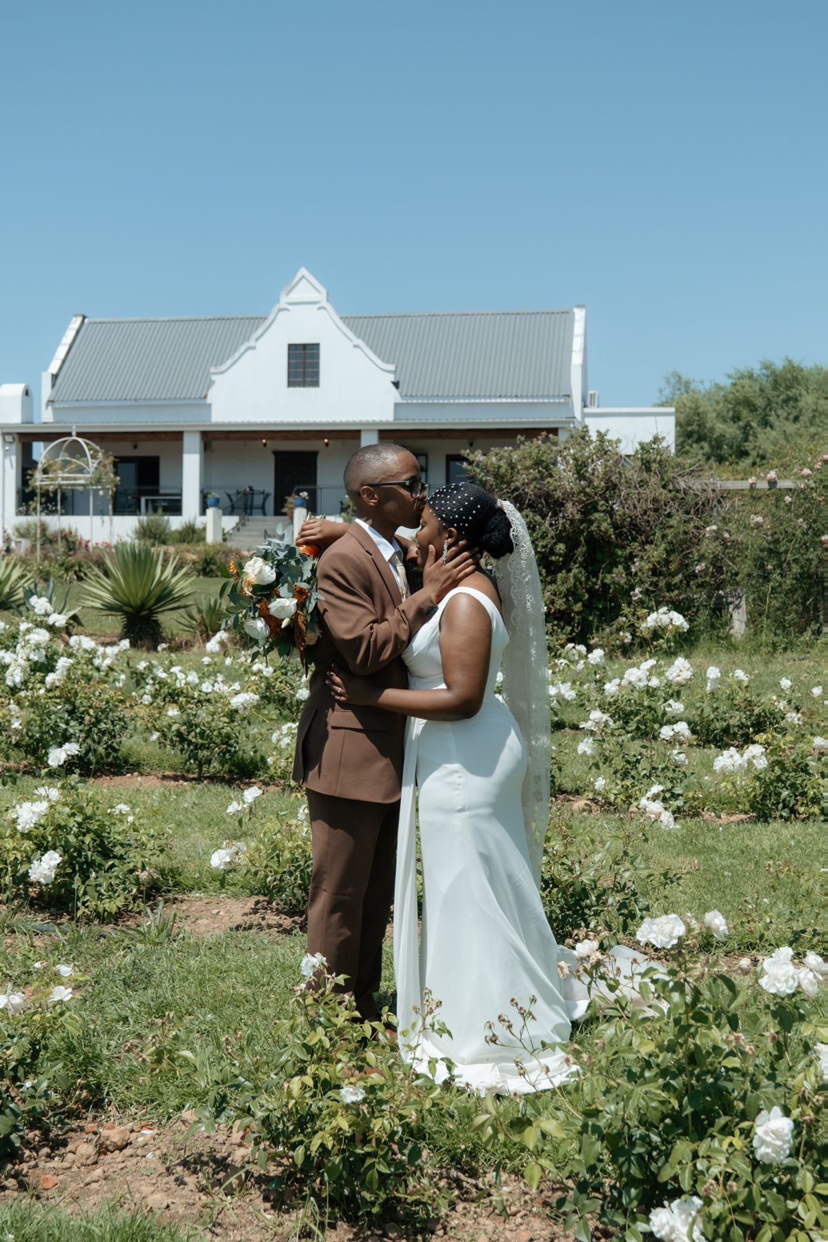 Black couple sharing a kiss in a white rose garden, bride in white gown with veil, groom in brown suit, Cape Dutch venue.