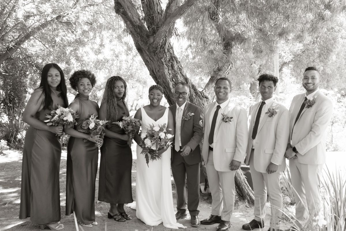 Black and white wedding party photo with bride, groom, bridesmaids and groomsmen posing under a tree outdoors.