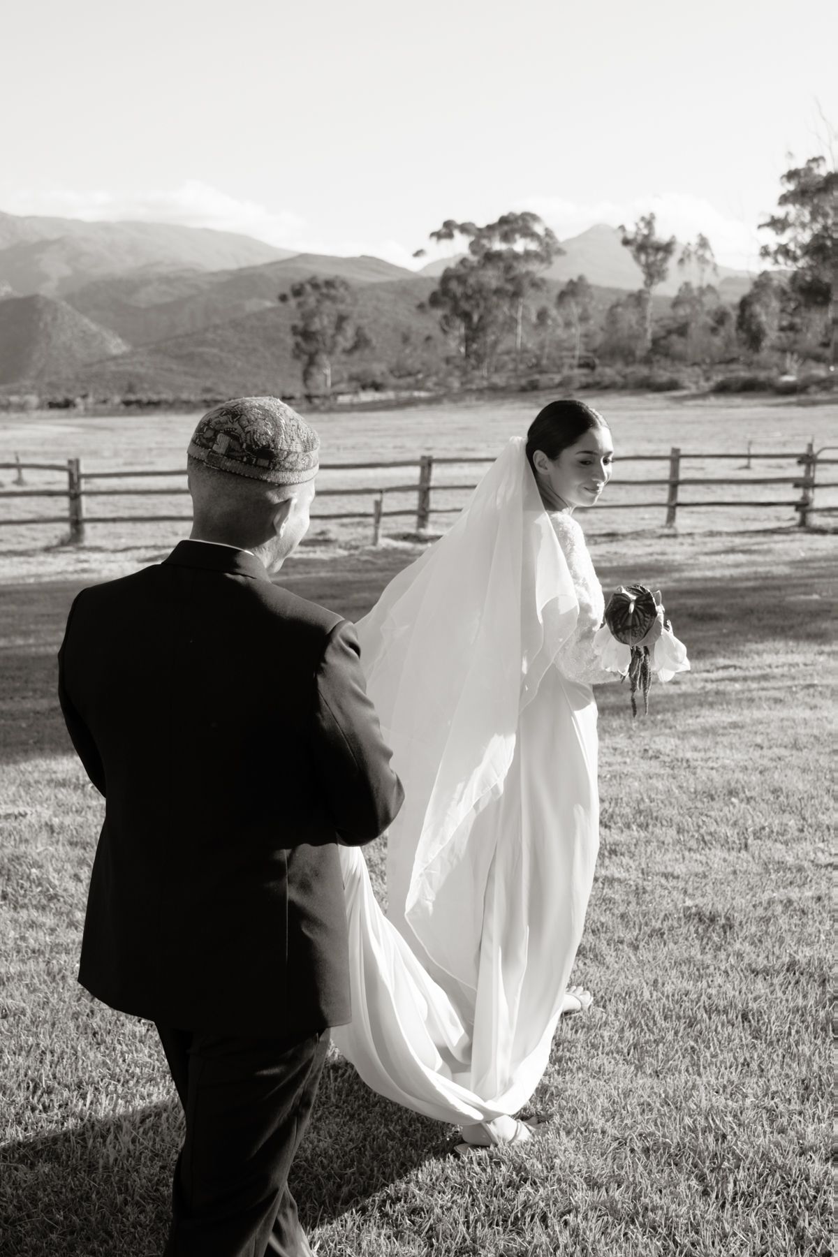 Black and white photo of groom holding bride's veil train in field with mountain backdrop and wooden fence.