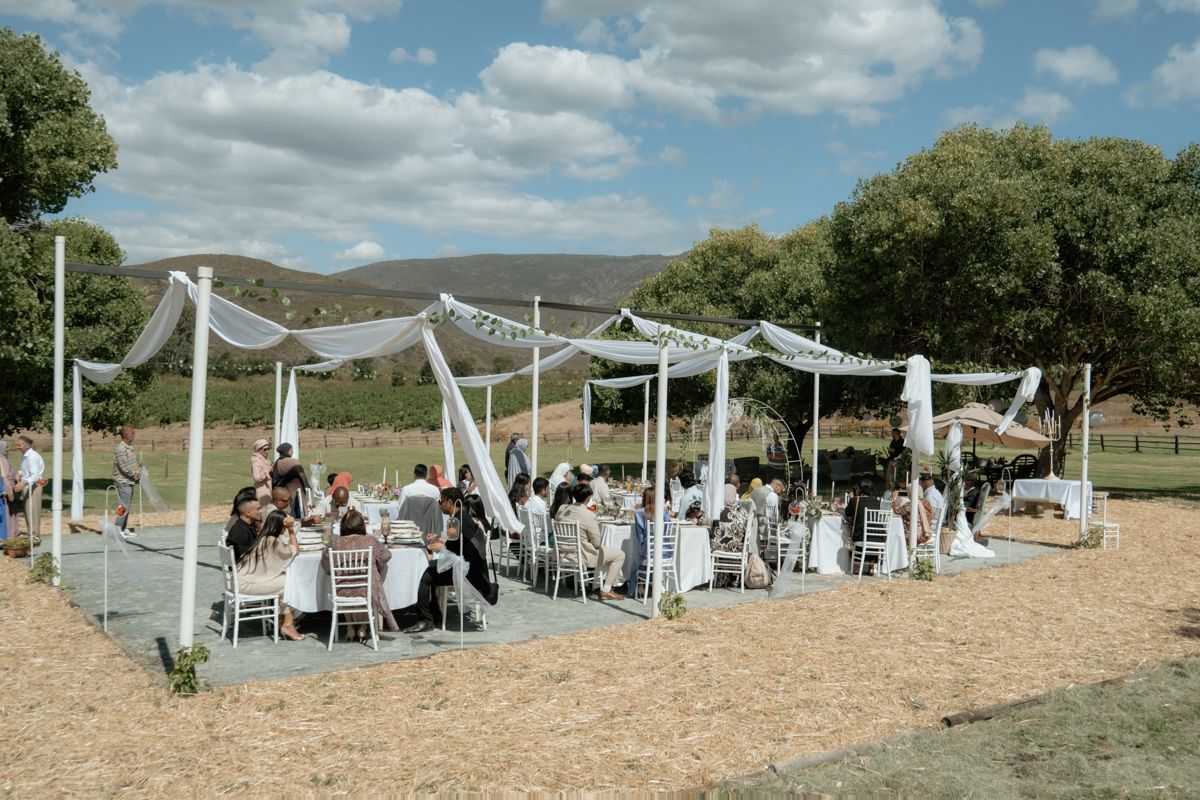 Outdoor wedding reception under white draped canopy with guests seated at tables in scenic countryside.
