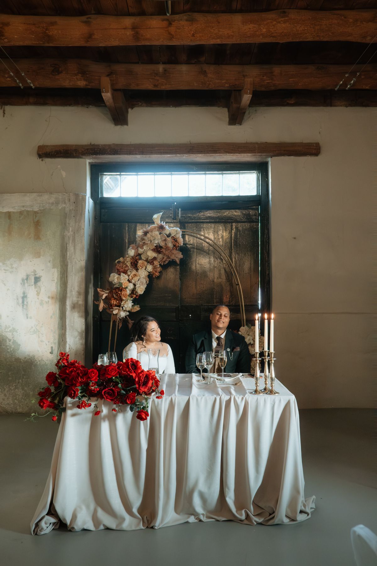 Bride and groom seated at sweetheart table adorned with red roses and candles beneath rustic wooden ceiling.