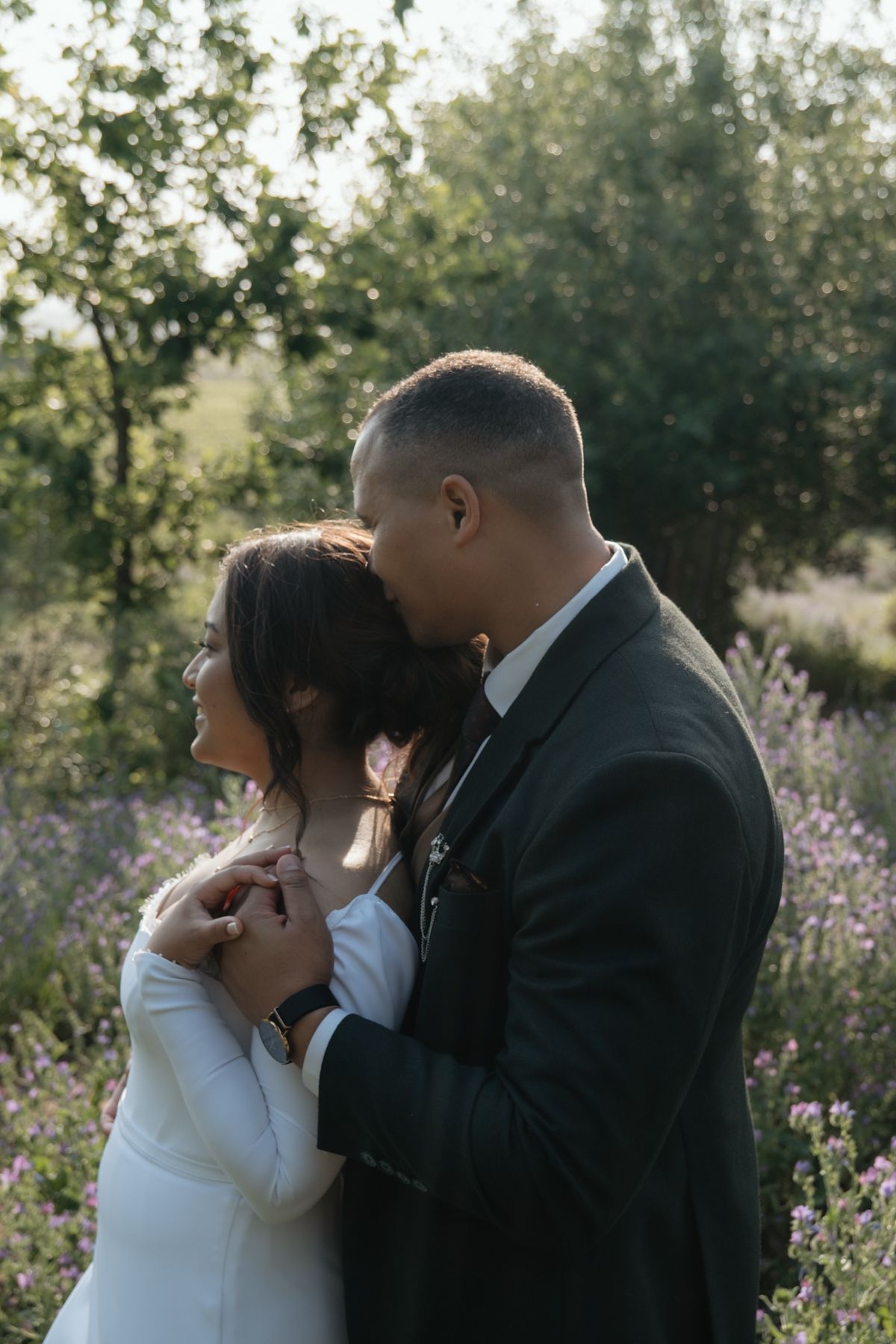 Groom in dark suit tenderly kissing bride on head in lavender field at golden hour.