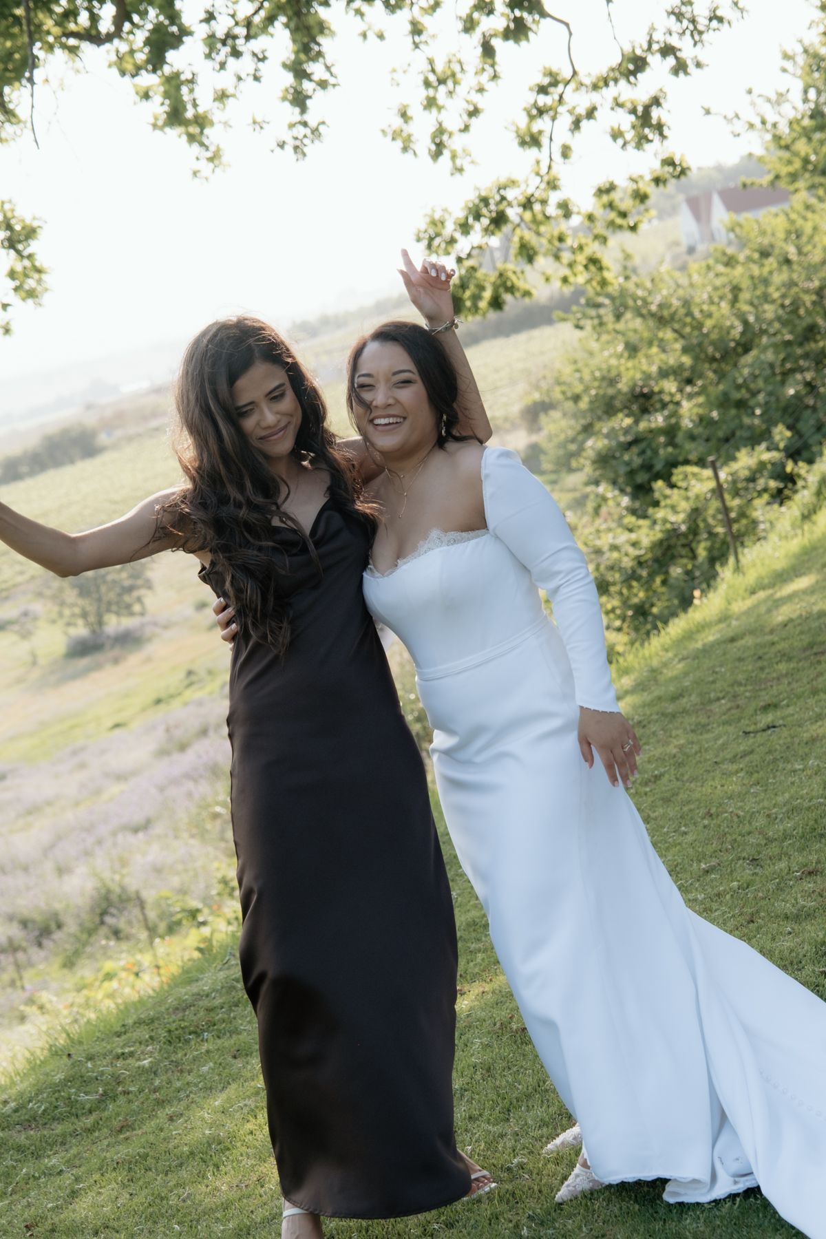 Joyful bride in white one-shoulder gown posing with bridesmaid in black dress on green hillside.