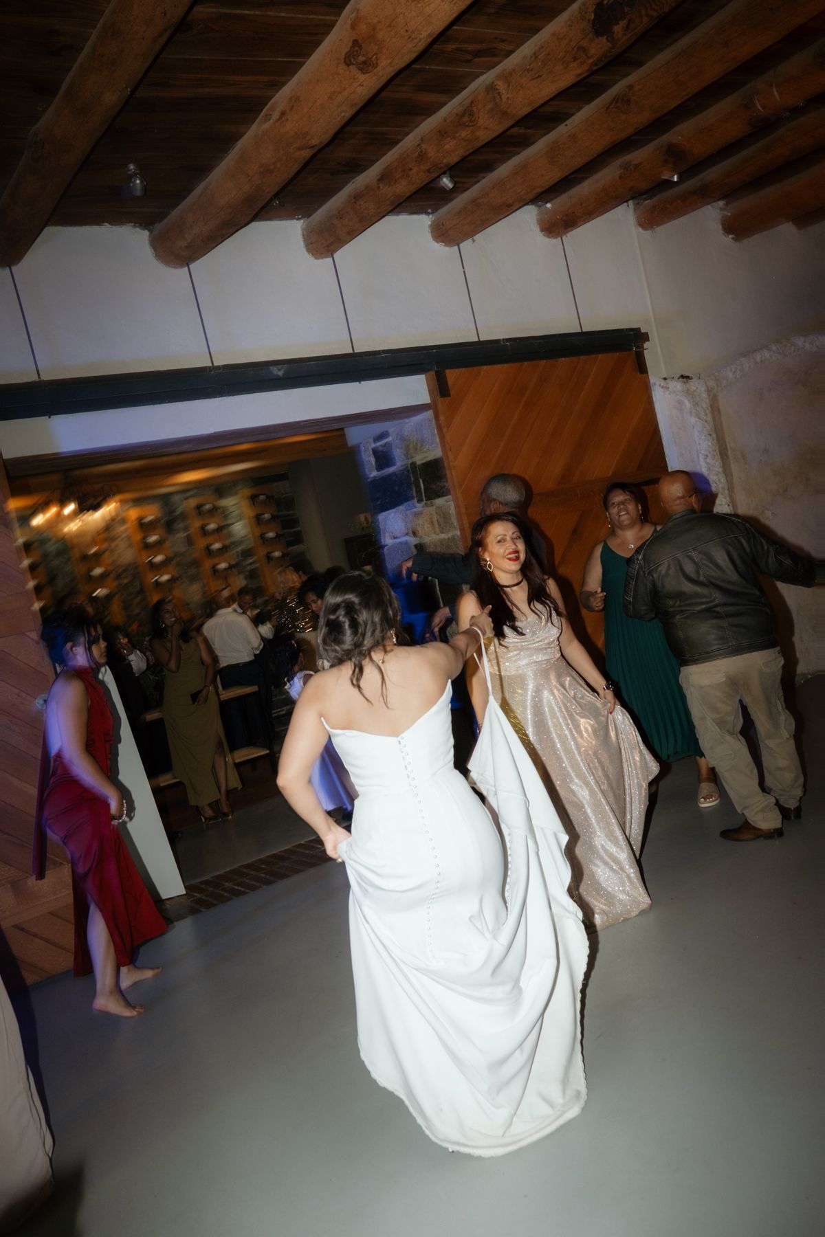 Bride in white strapless gown dances with guests on the dance floor at a rustic wedding reception.