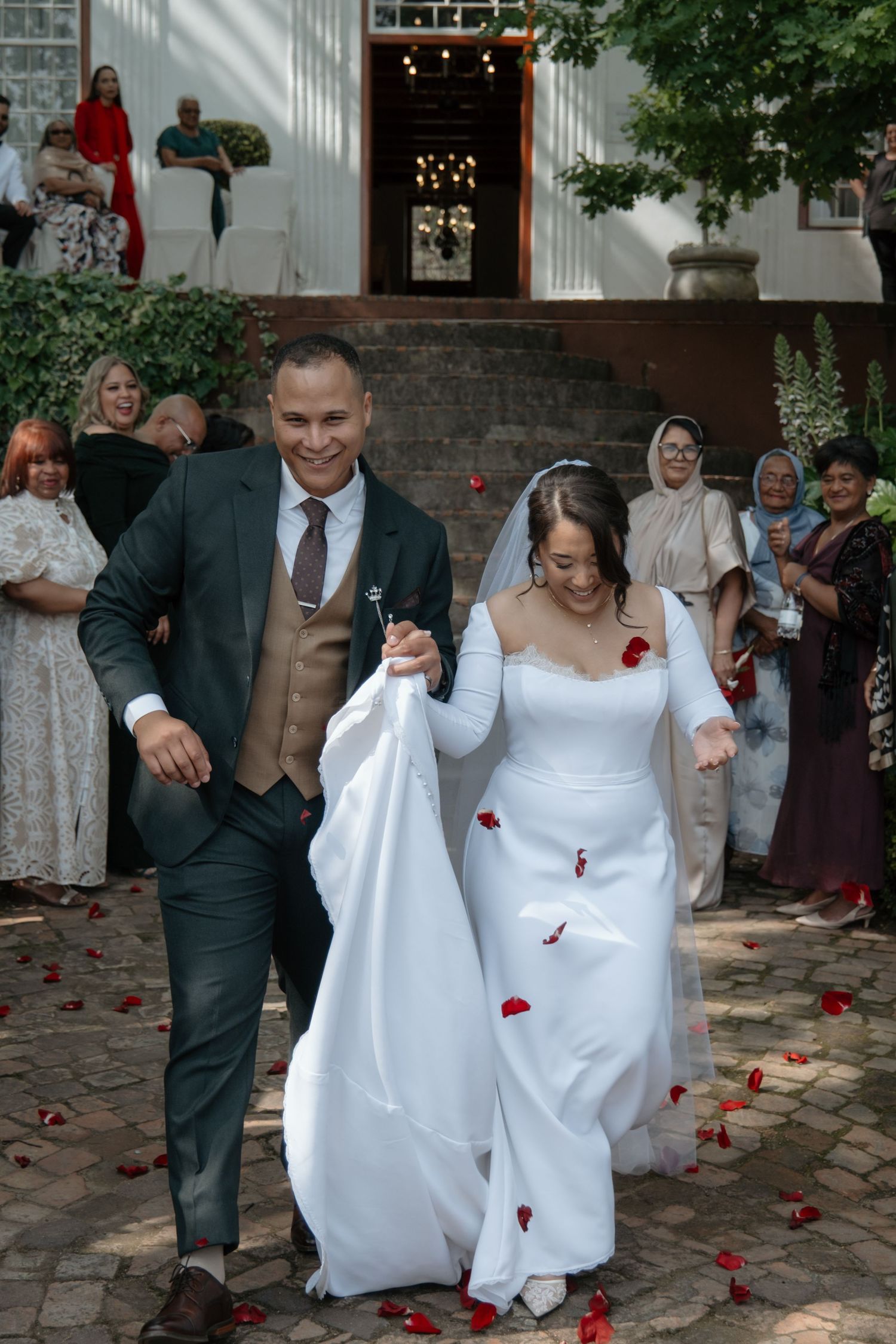 Joyful newlywed couple walking through rose petal confetti as wedding guests celebrate outside a historic venue.
