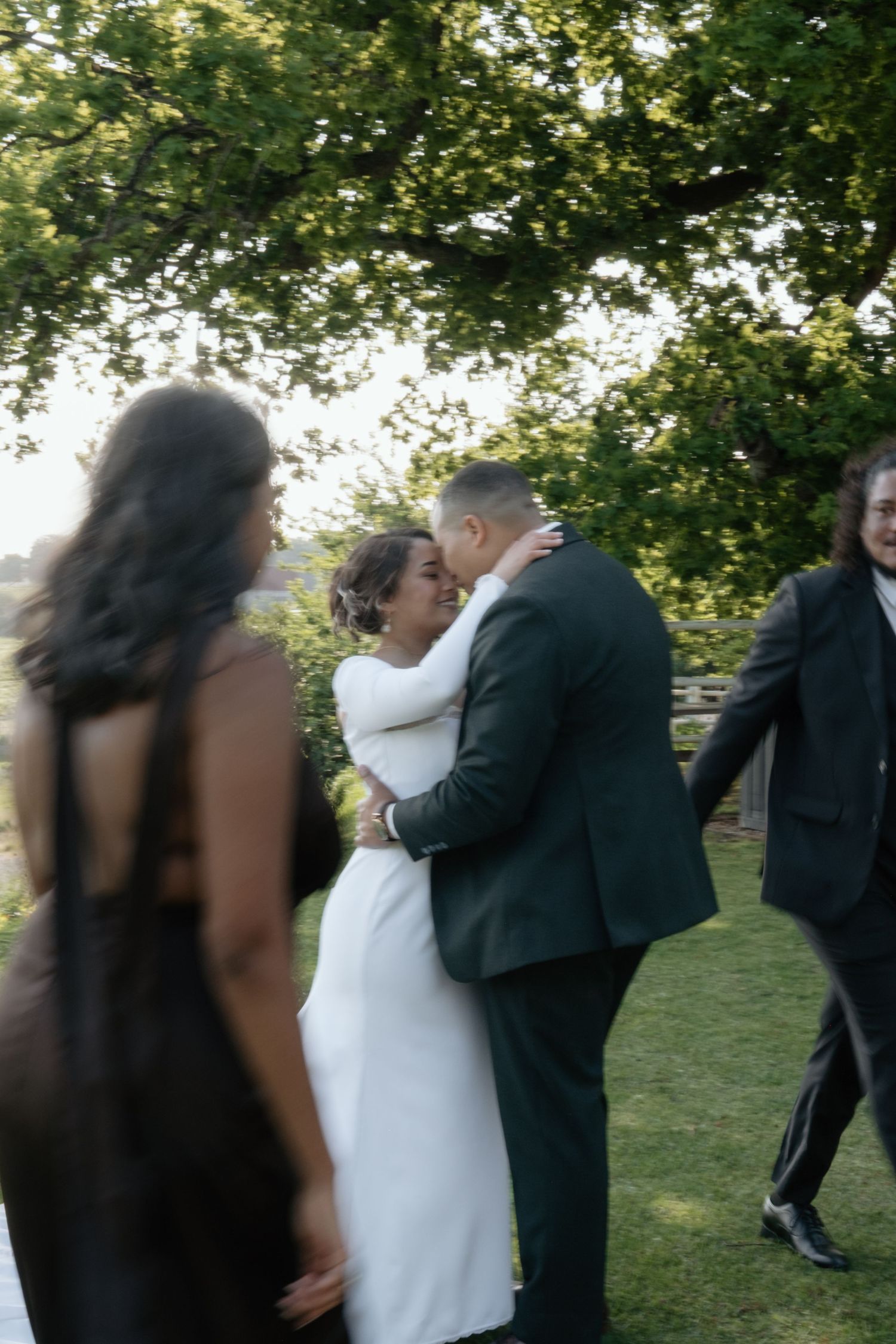 Bride and groom share a romantic kiss outdoors at their wedding reception under a green tree.
