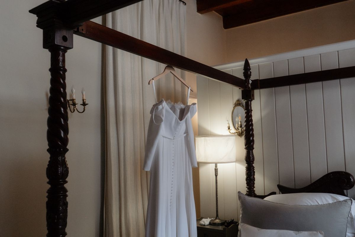 Elegant white wedding dress with long sleeves hanging on a dark wood four-poster bed frame.