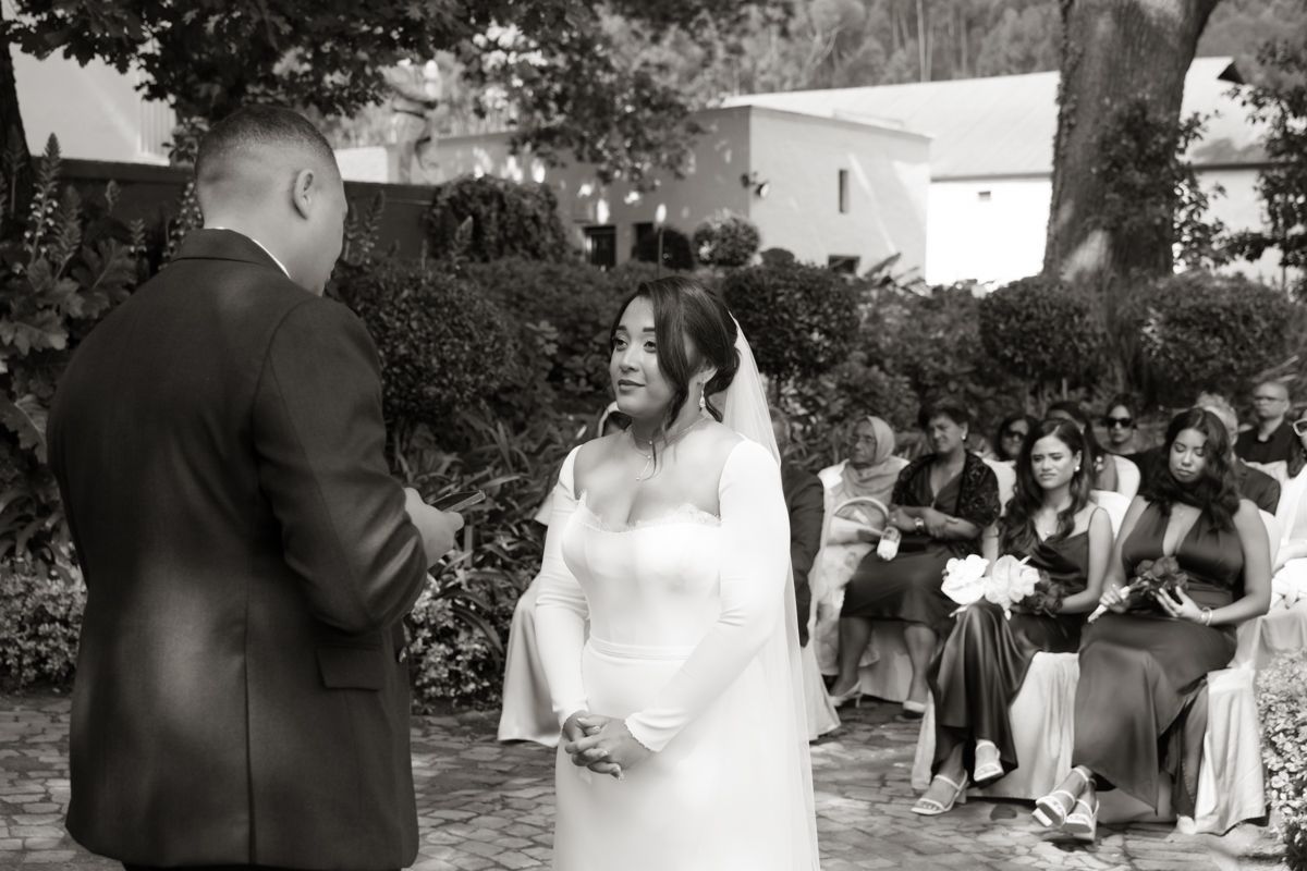 Black and white photo of a bride in a white gown and veil exchanging vows with the groom at an outdoor ceremony.