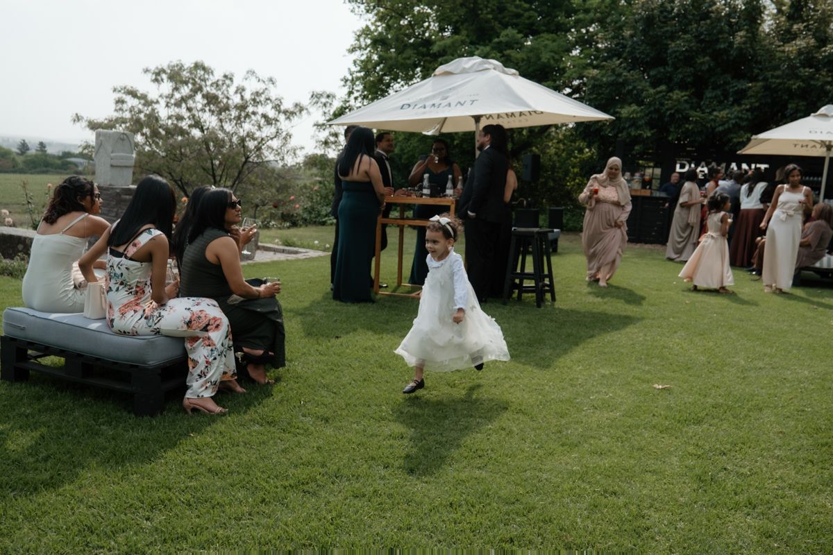 A flower girl in white dress runs across green lawn at an outdoor wedding reception with guests seated nearby.