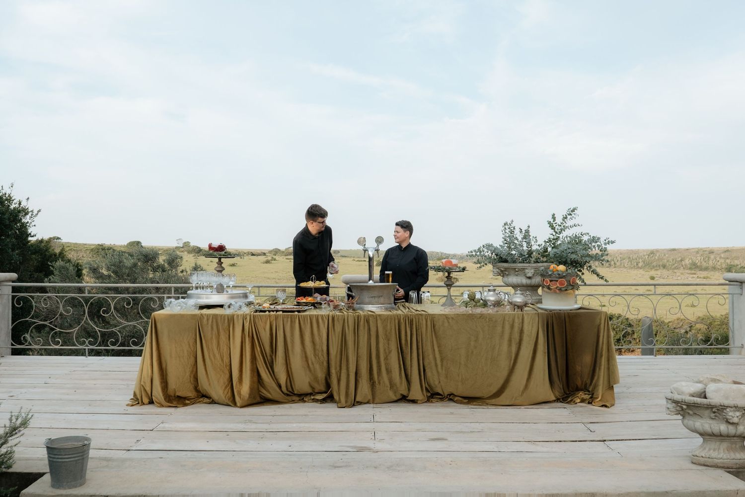 Two catering staff in black uniforms standing behind a gold-draped buffet table at outdoor event.