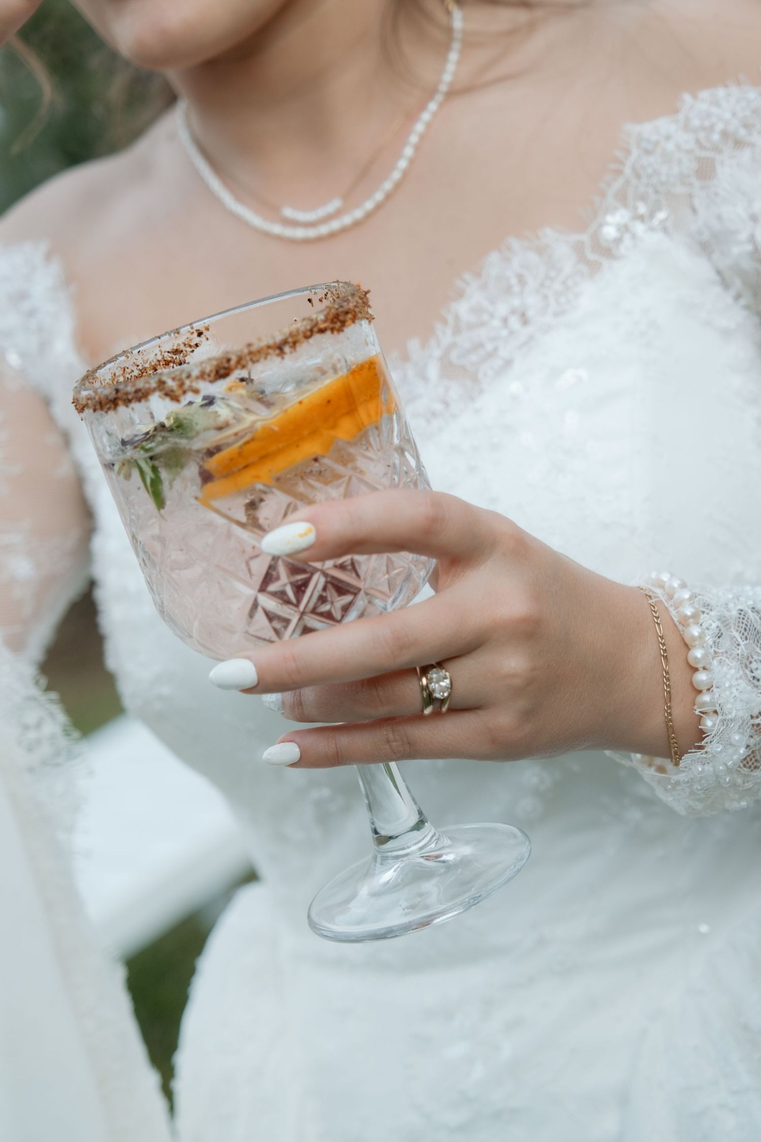 Close-up of bride holding elegant cocktail glass with orange garnish, wearing diamond engagement ring.
