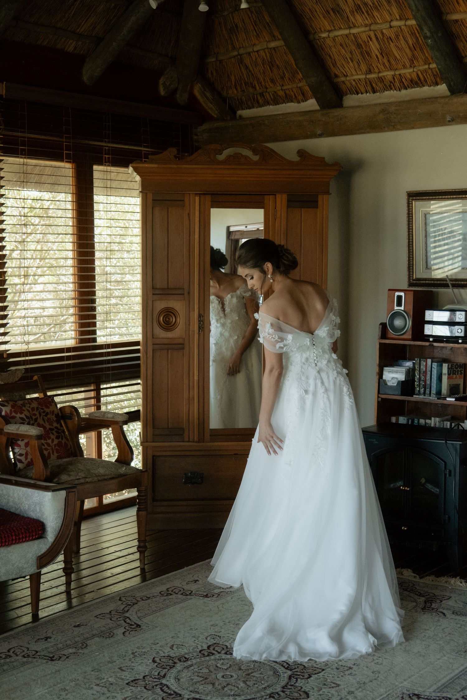 Bride in off-shoulder white gown admires her reflection in a wooden wardrobe mirror inside a rustic lodge room.