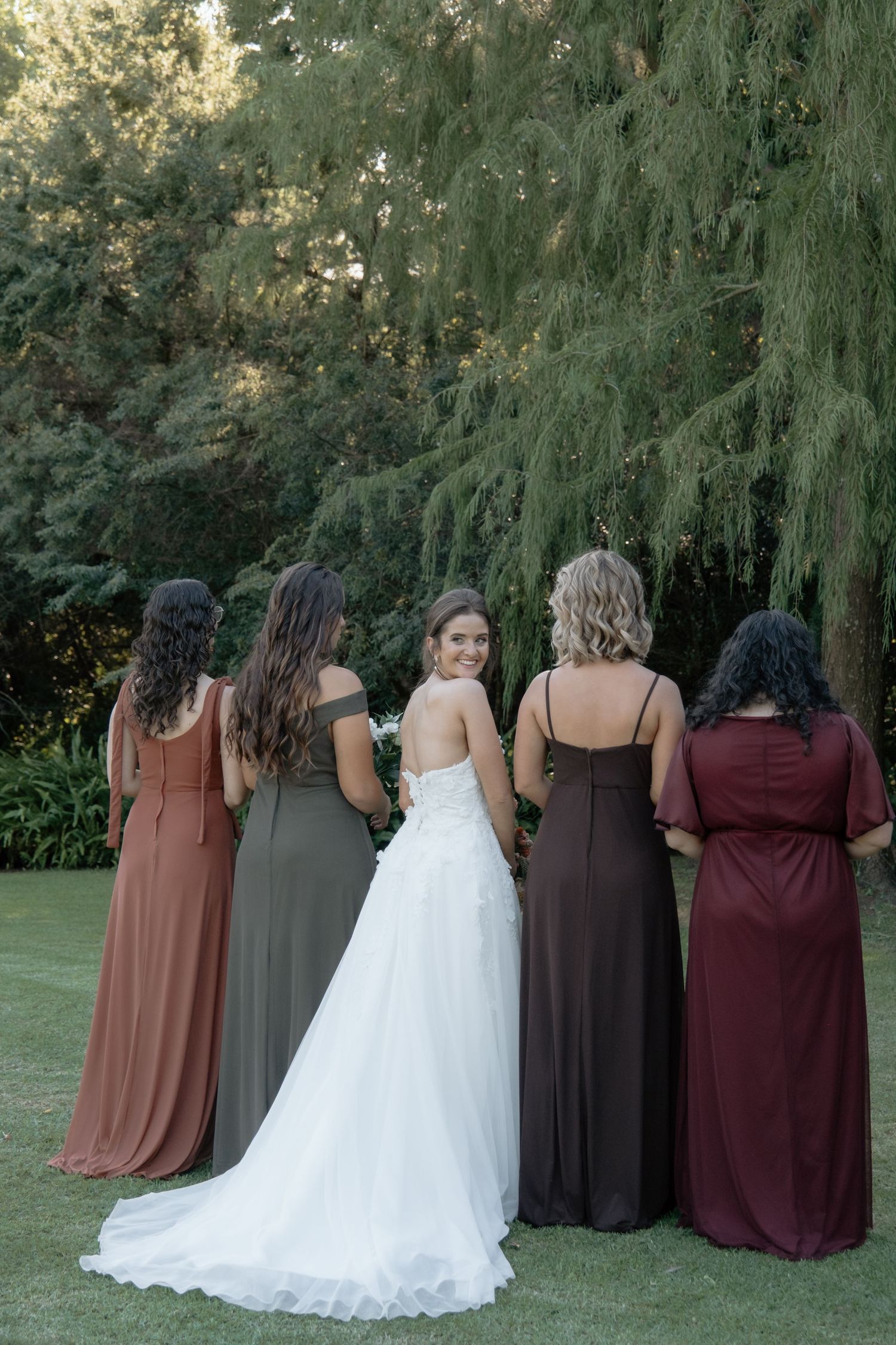 Bride in white gown turns to smile while standing with bridesmaids in earthy-toned dresses outdoors.