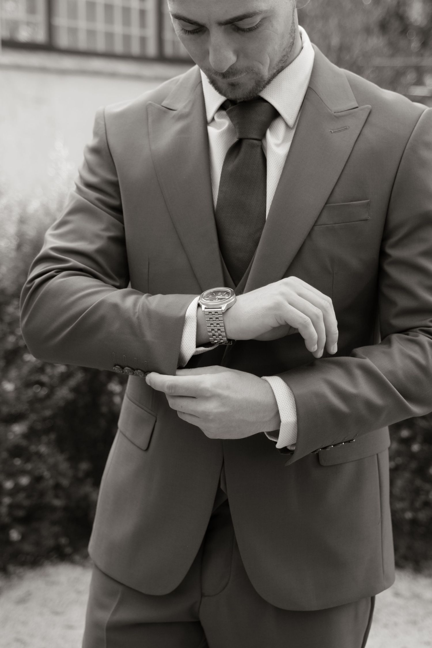 Groom in a grey suit adjusting his watch cufflinks before wedding ceremony, black and white photo.