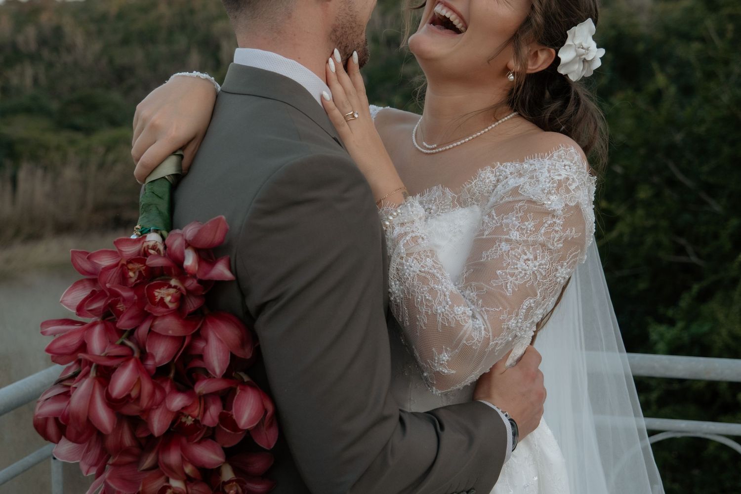 Joyful bride in lace gown laughs with groom while holding a bouquet of deep red calla lilies outdoors.