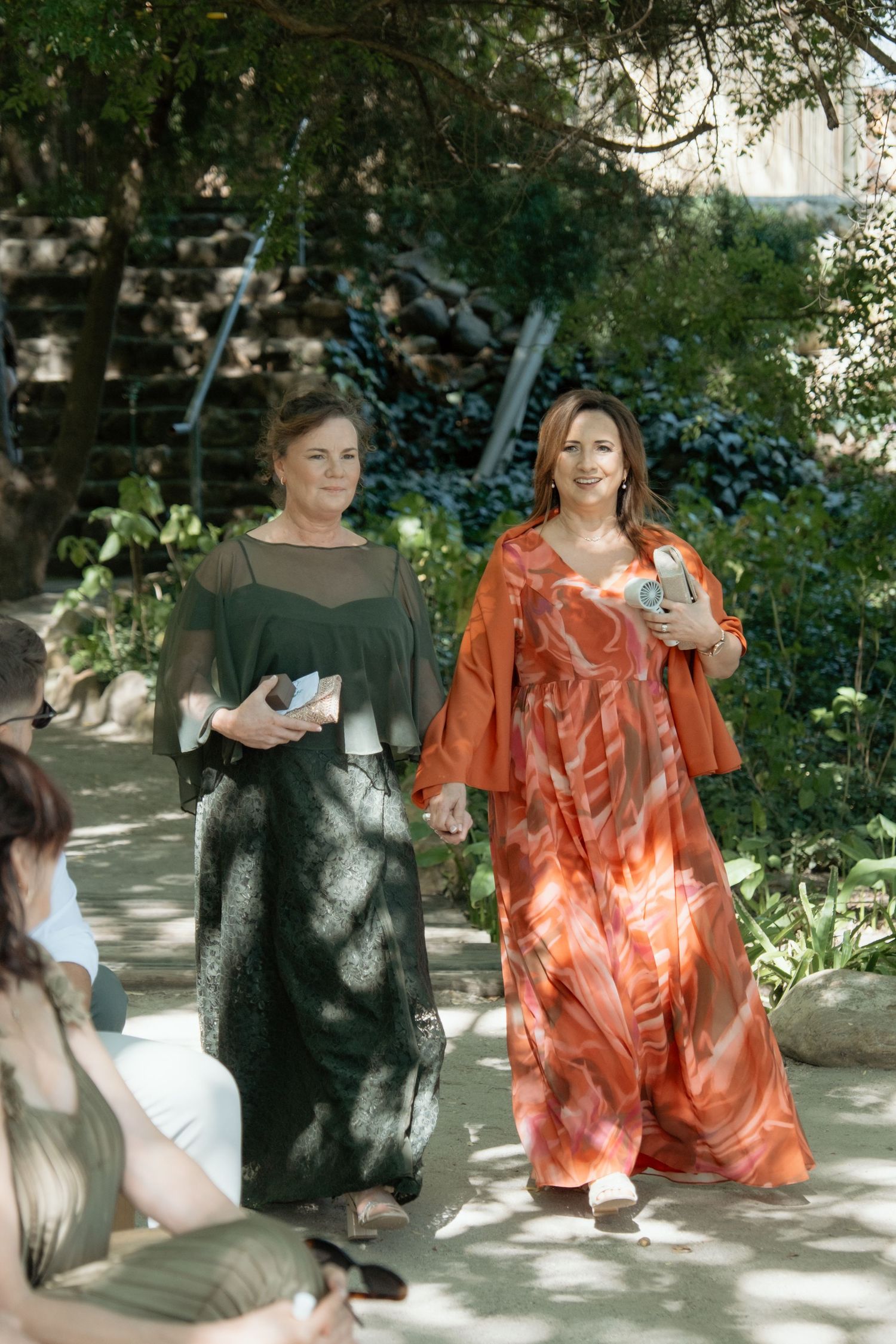 Two women in elegant green and orange dresses walking down the aisle at an outdoor garden wedding ceremony.
