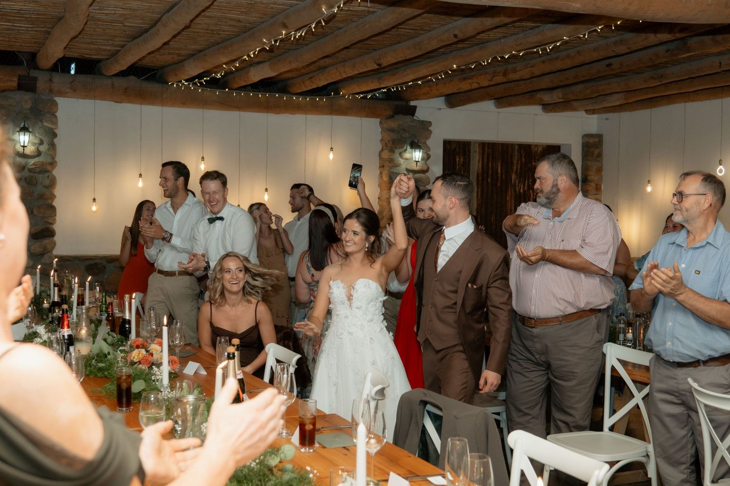 Bride and groom make a grand entrance into their rustic wedding reception as guests cheer and celebrate.
