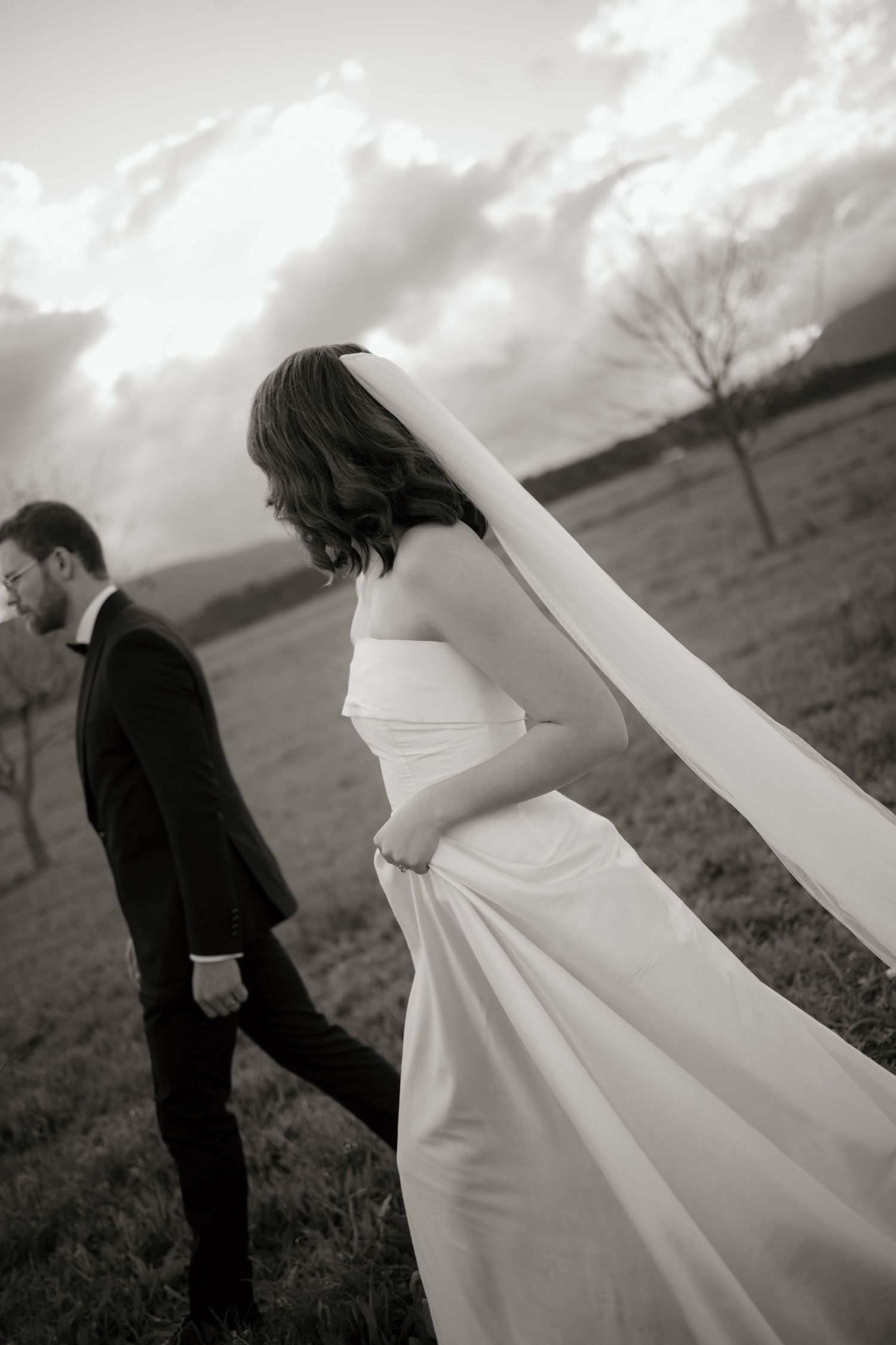 Black and white photo of bride in flowing gown and veil walking with groom in suit across a field.