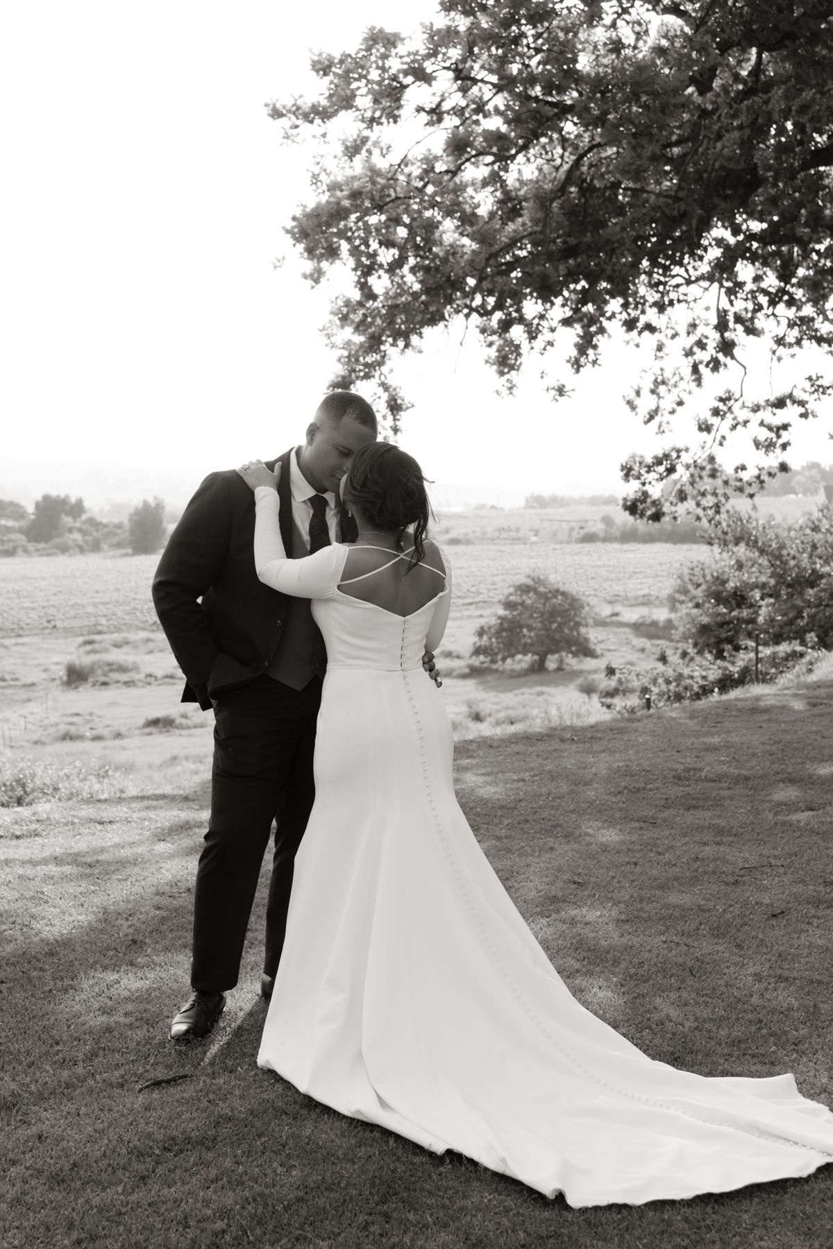 Bride and groom share a romantic kiss in black and white photo beneath a tree at outdoor wedding.