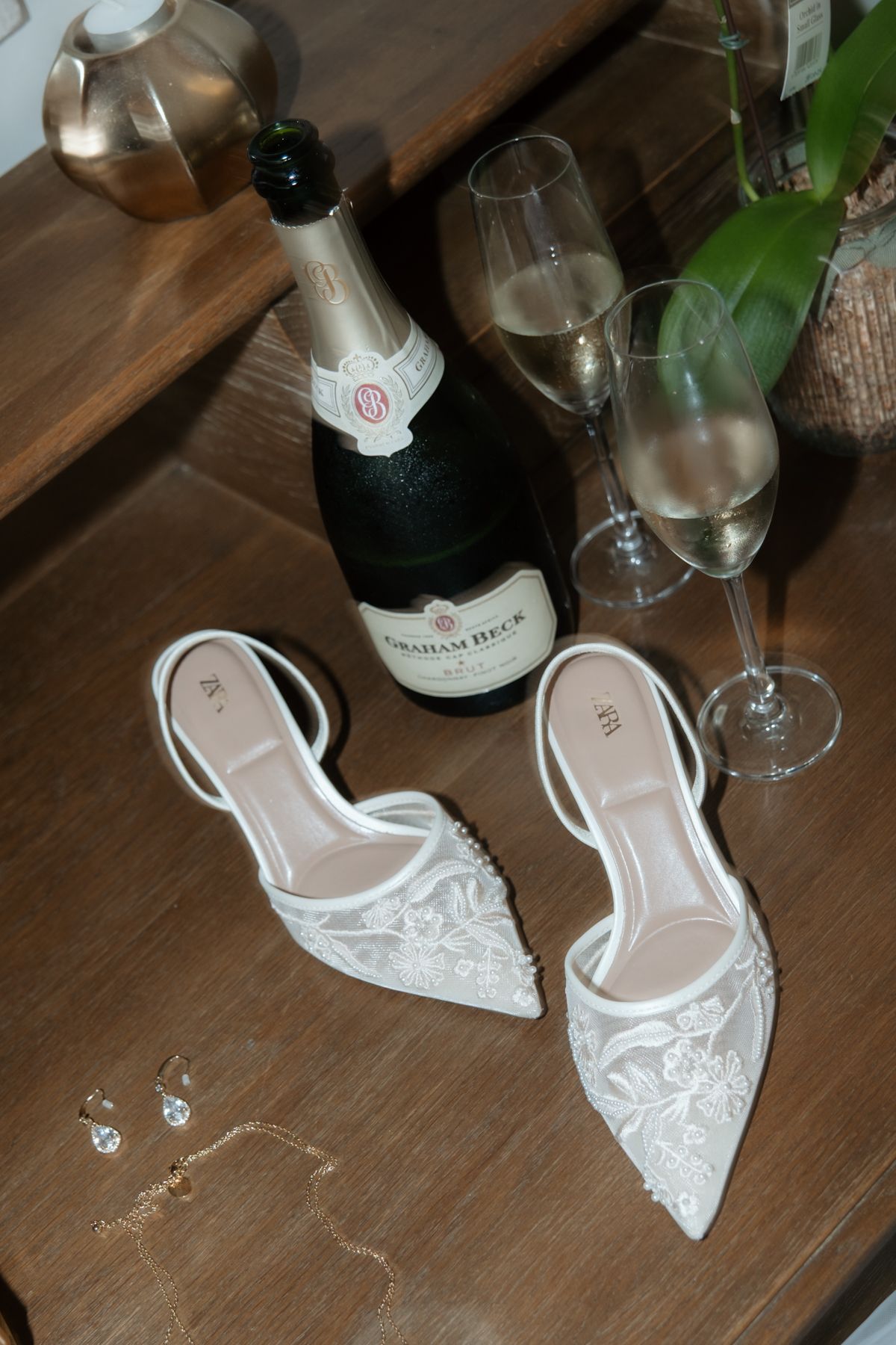 White lace bridal heels beside champagne bottle and glasses on wooden table.