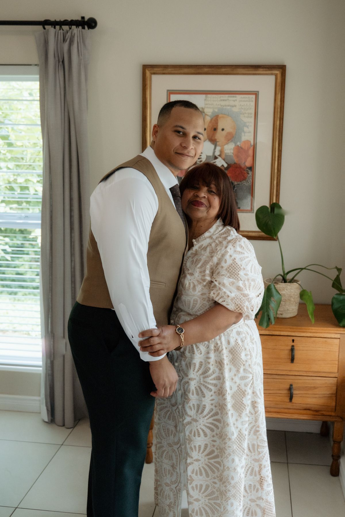 Groom in dark suit and tan vest posing with his mother in a white lace dress at home.