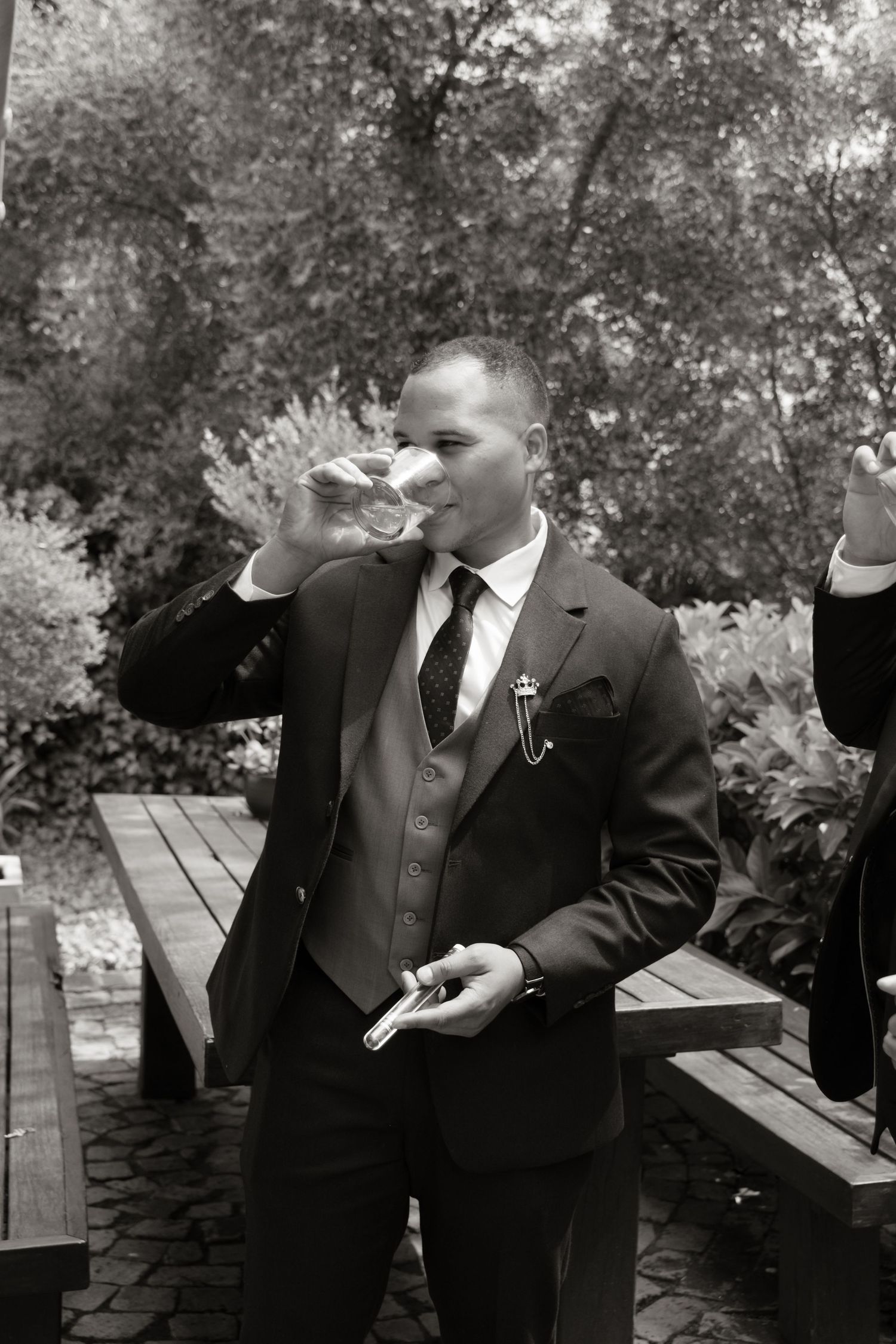 Groom in suit drinking champagne while holding a cigar outdoors at wedding celebration.