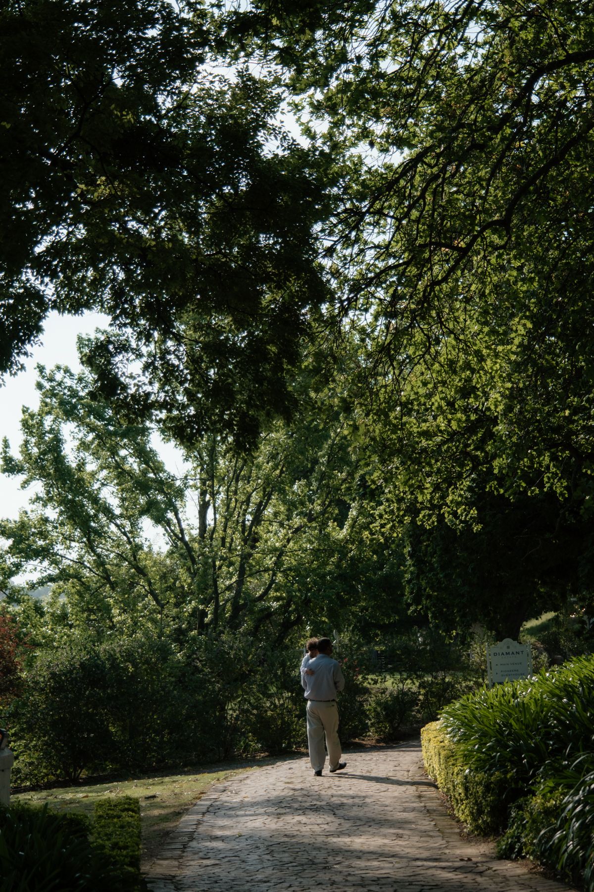 A lone guest walks along a sunlit stone pathway surrounded by lush green trees at a garden wedding venue.
