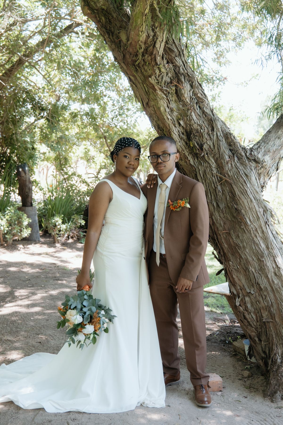 Bride in white gown and groom in brown suit pose together under a tree holding an orange and white bouquet.