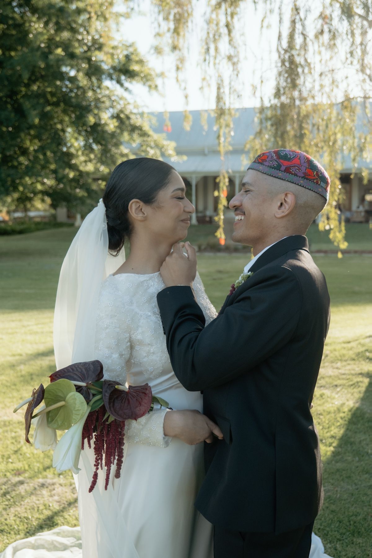 Smiling bride and groom in intimate embrace on green lawn, groom wearing decorative kufi cap.