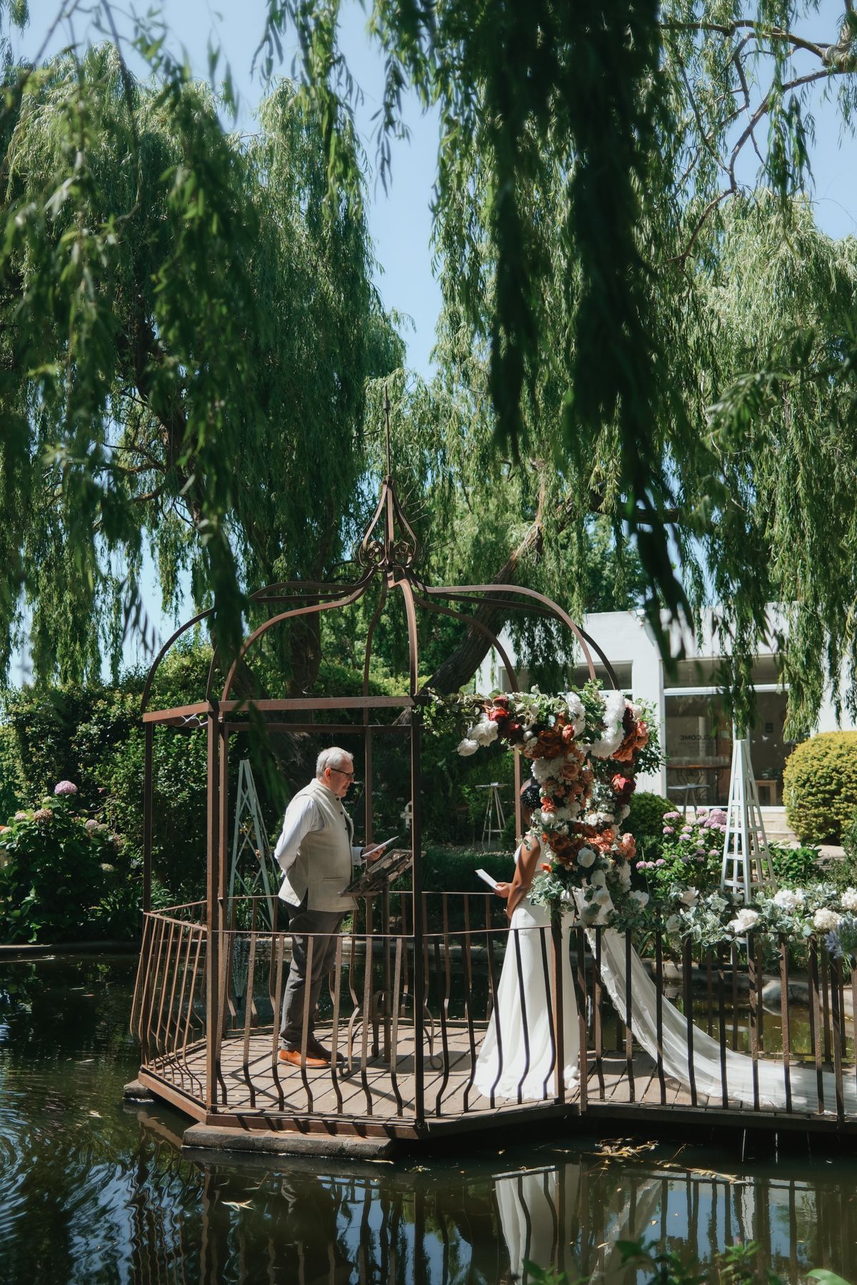 Officiant and bride stand on rustic iron gazebo over pond at garden wedding.