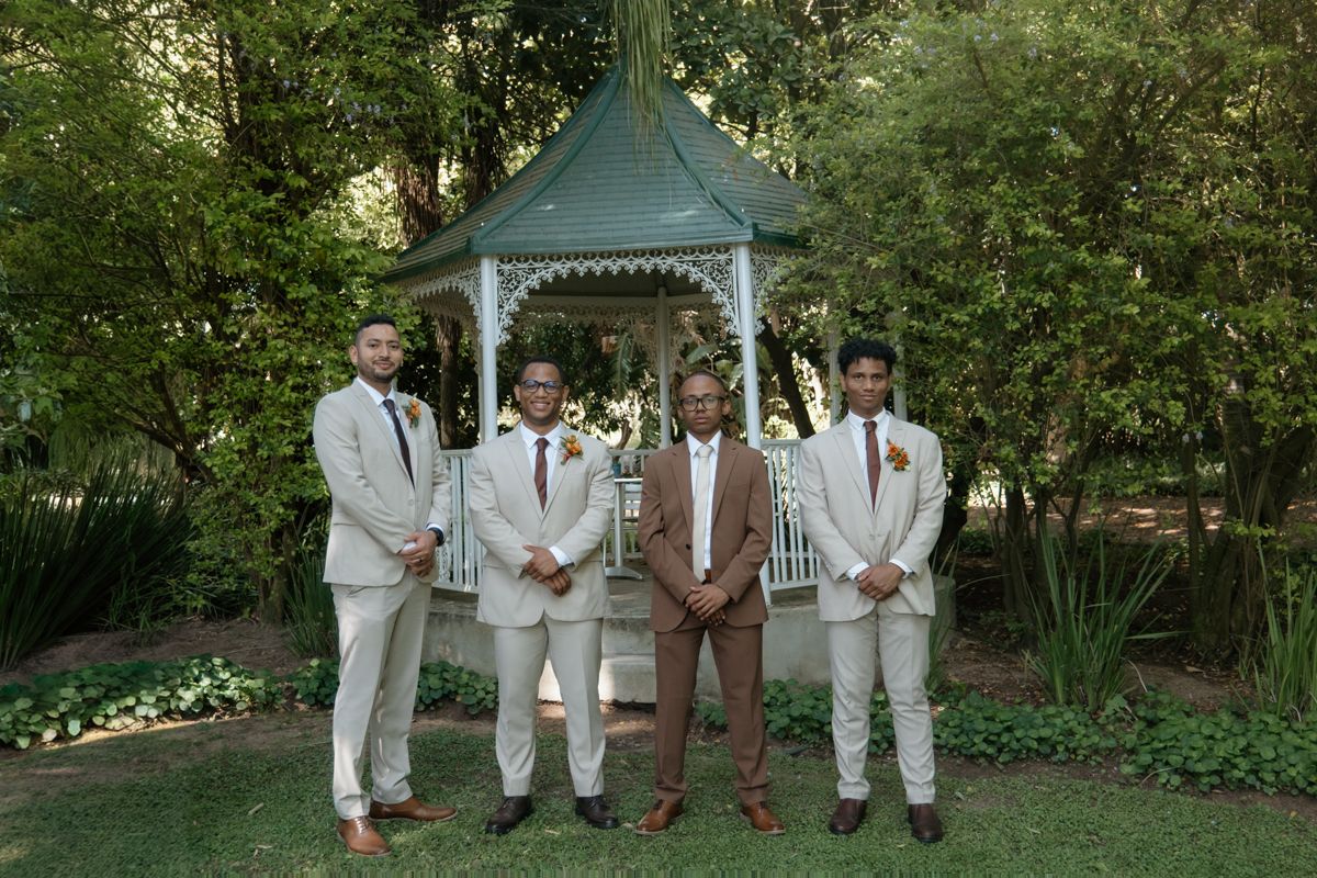 Four groomsmen in tan and brown suits posing in front of a white gazebo surrounded by lush green trees.