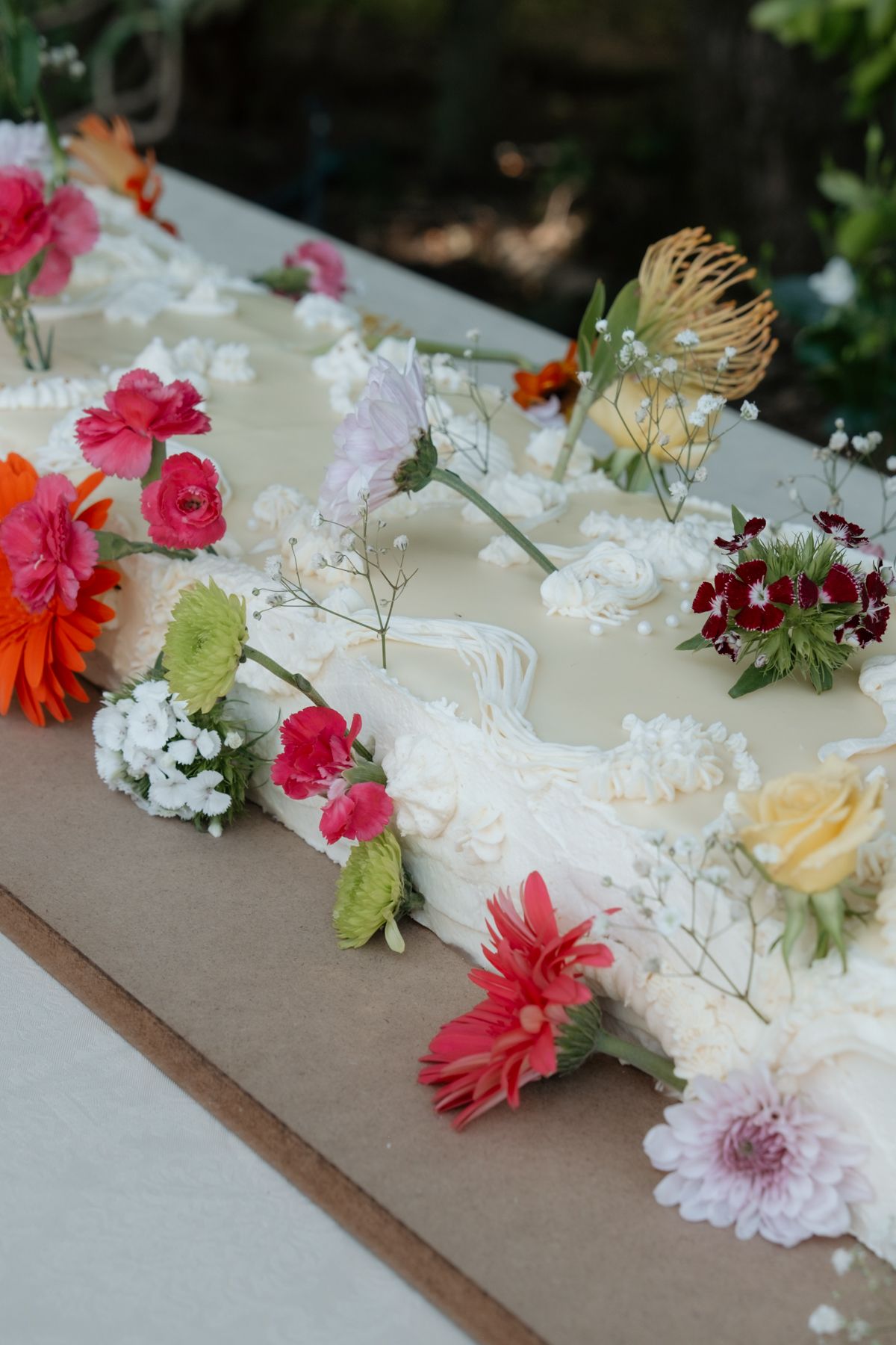 Elegant wedding cake decorated with colorful fresh flowers including pink gerberas, roses, and wildflowers on a white tablecloth.