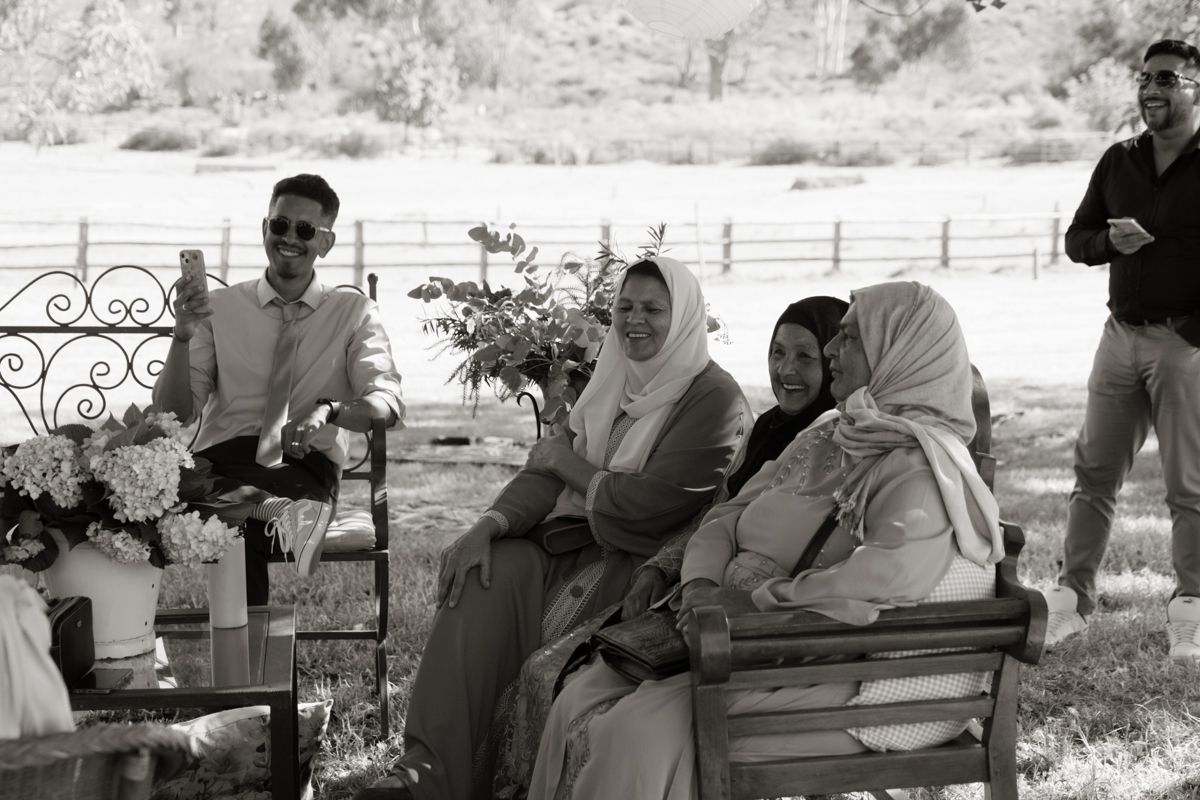 Black and white photo of wedding guests seated outdoors, women wearing hijabs at rural venue.