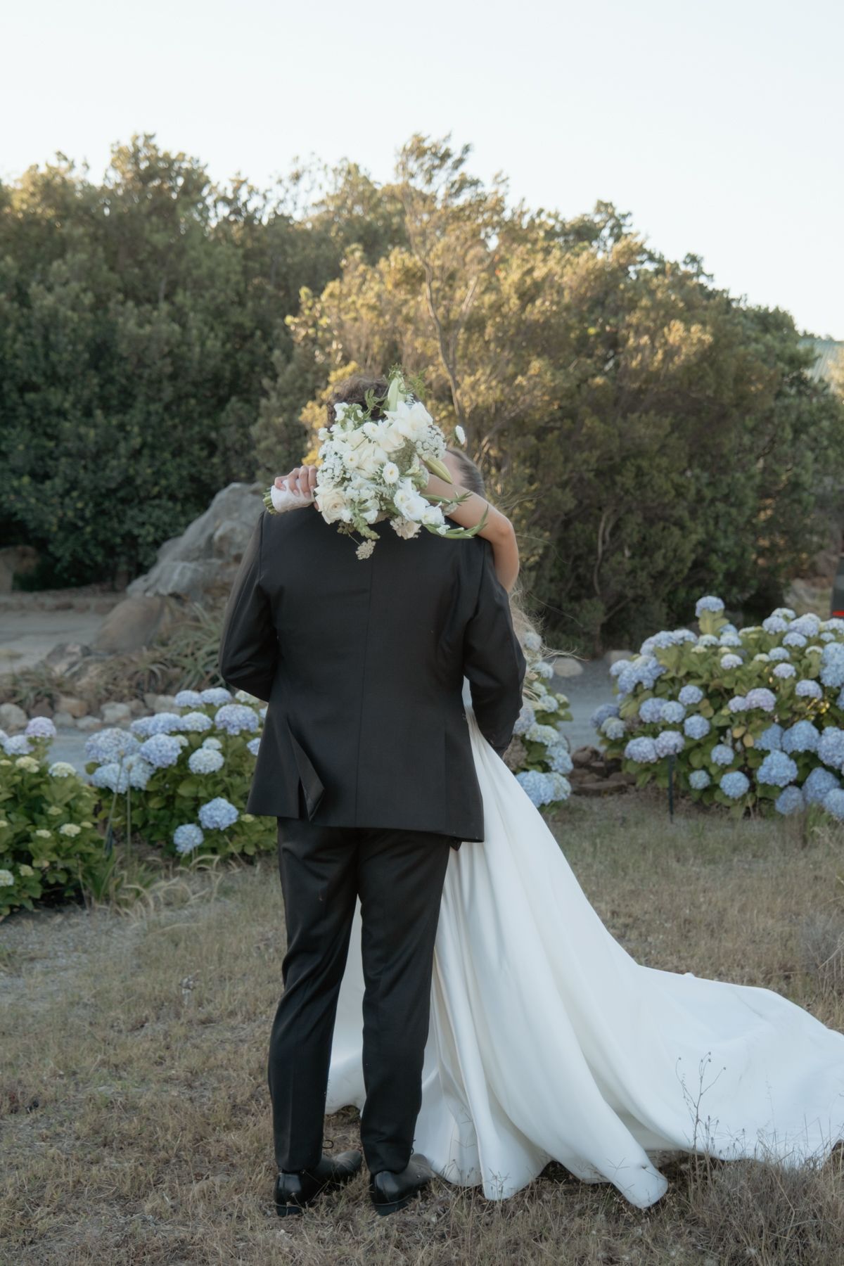 Bride and groom from behind sharing kiss with white floral bouquet among blue hydrangea garden.