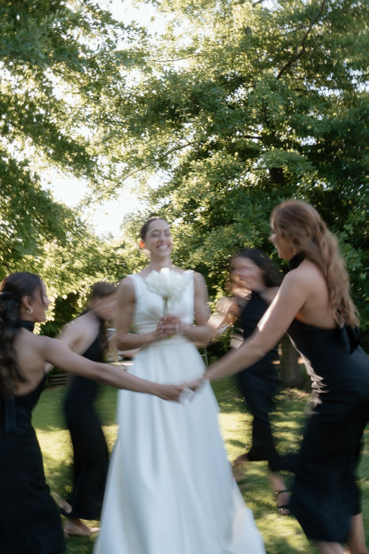 Smiling bride in white gown twirling with bridesmaids in black dresses outdoors beneath a canopy of green trees.