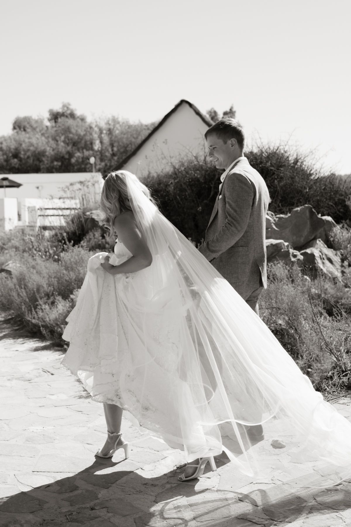 Black and white photo of bride and groom walking along stone path at outdoor wedding venue.