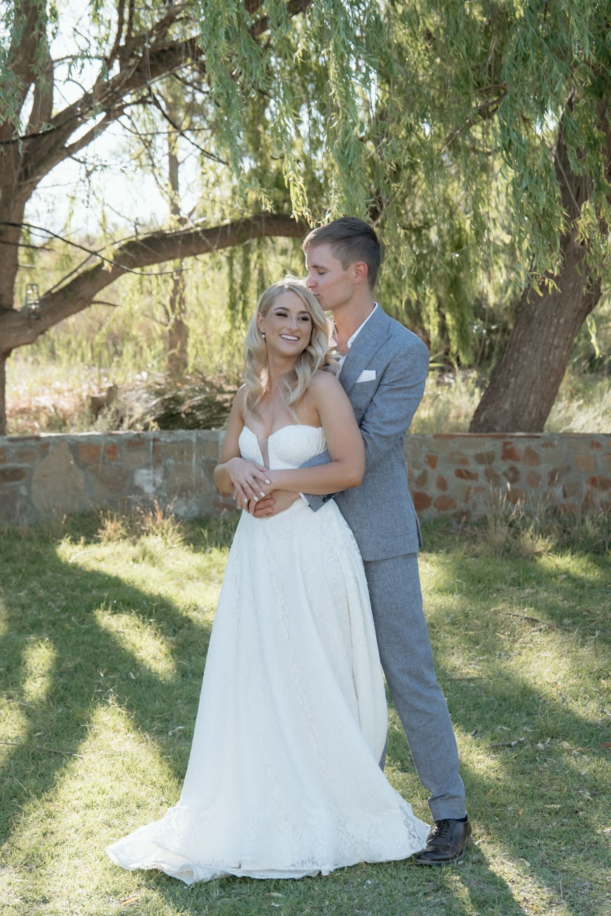 Groom in grey suit embracing bride in strapless white gown while kissing her cheek under a willow tree.