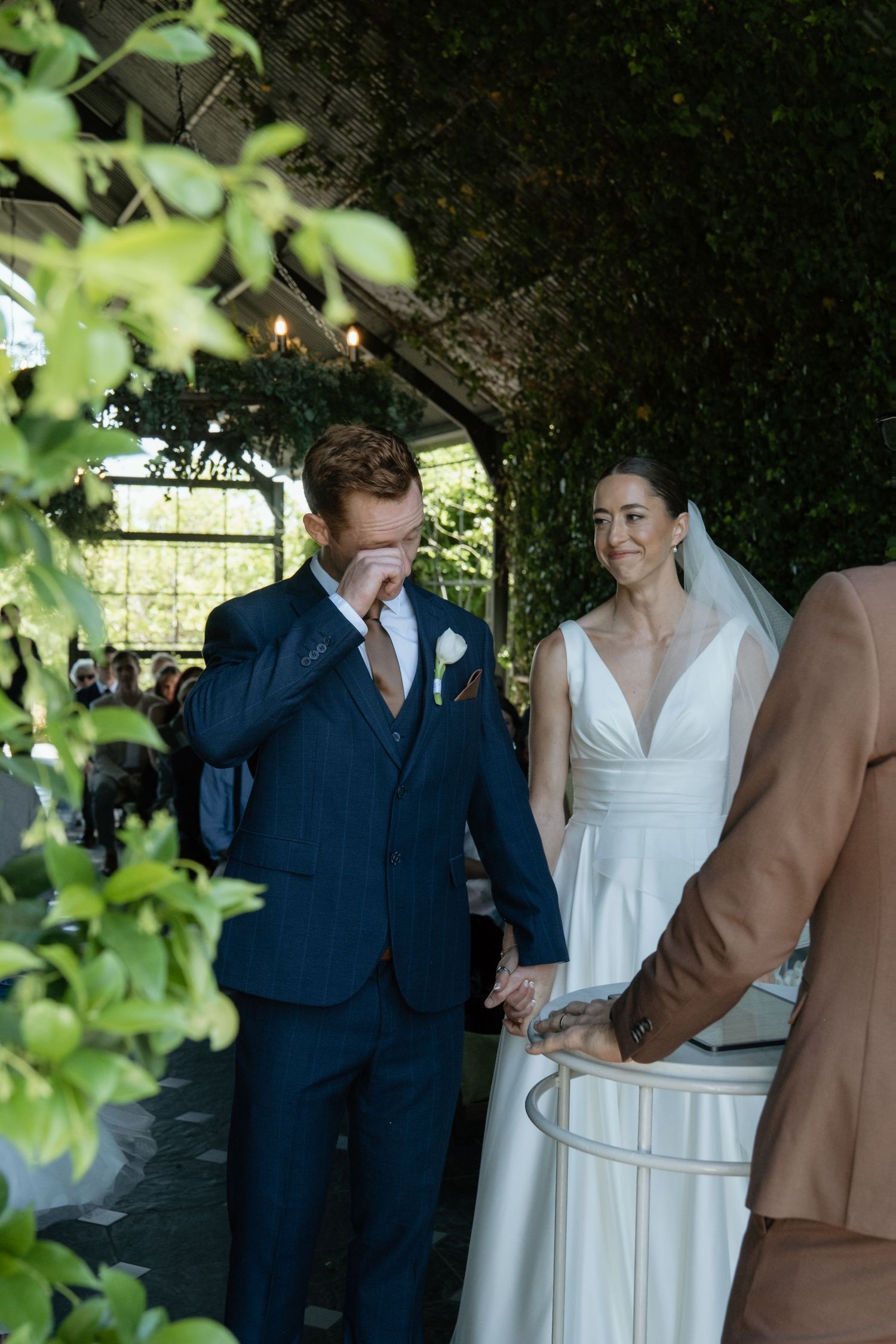 Emotional groom wipes tears holding brides hand during heartfelt outdoor wedding ceremony.