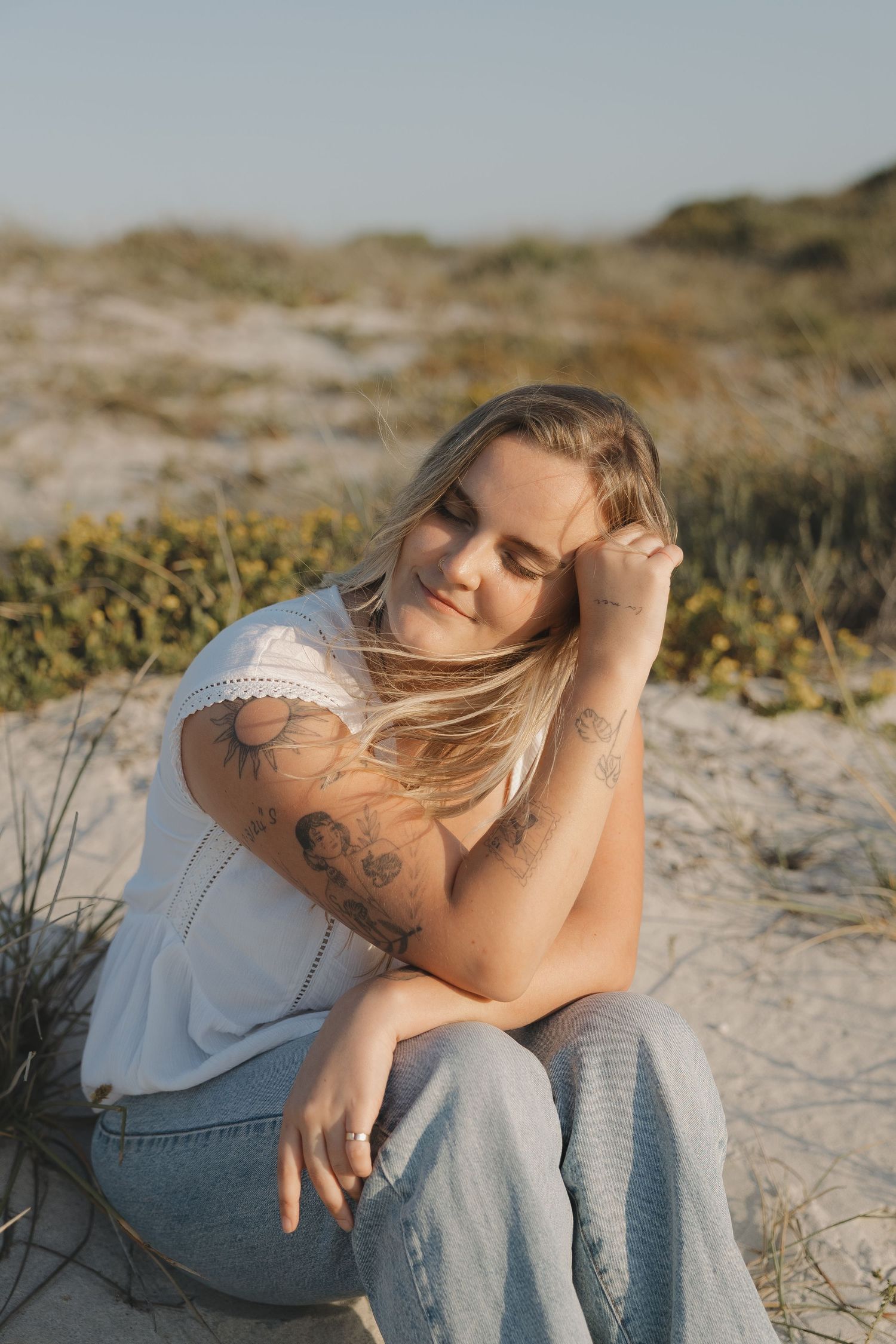 A person in a white top sits on sandy dunes with wind-blown hair during a golden hour photoshoot.