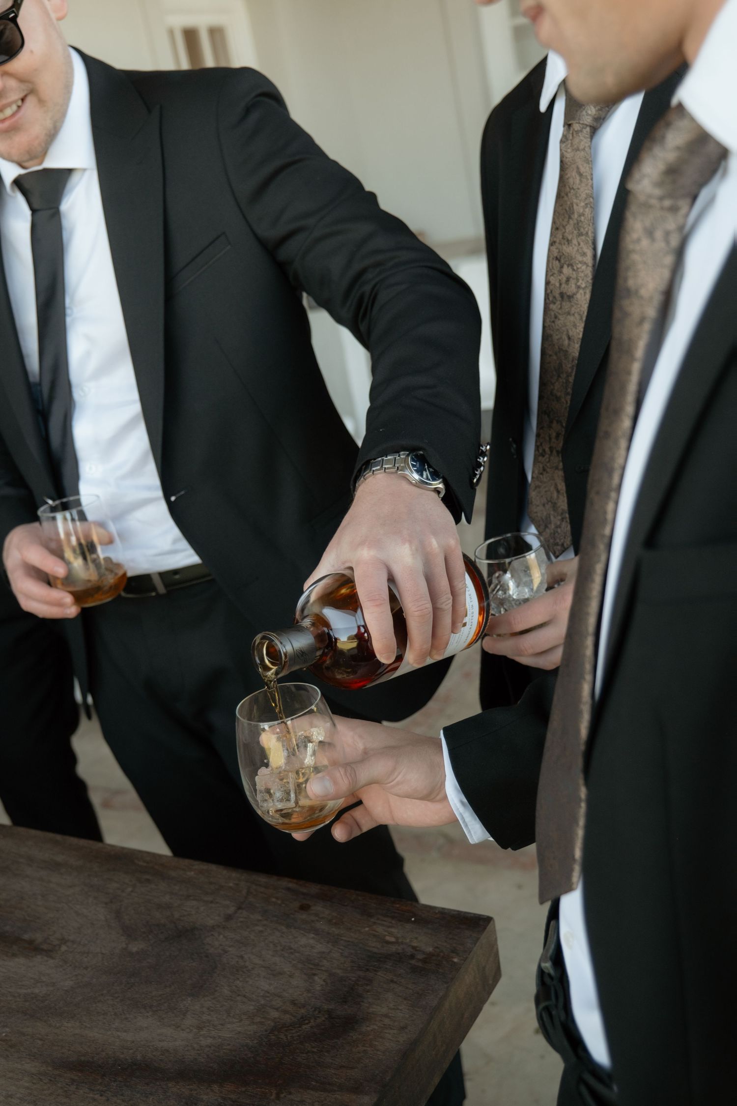 Men in black suits pouring whiskey into glasses with ice during a celebration or bachelor party.
