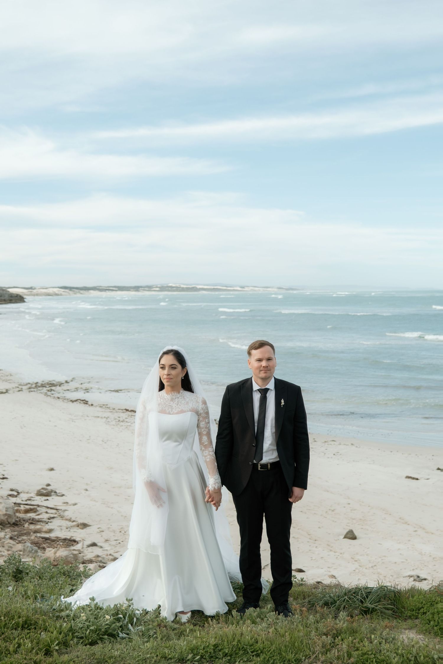 Bride and groom hold hands on sandy beach with ocean waves and cloudy sky behind them.