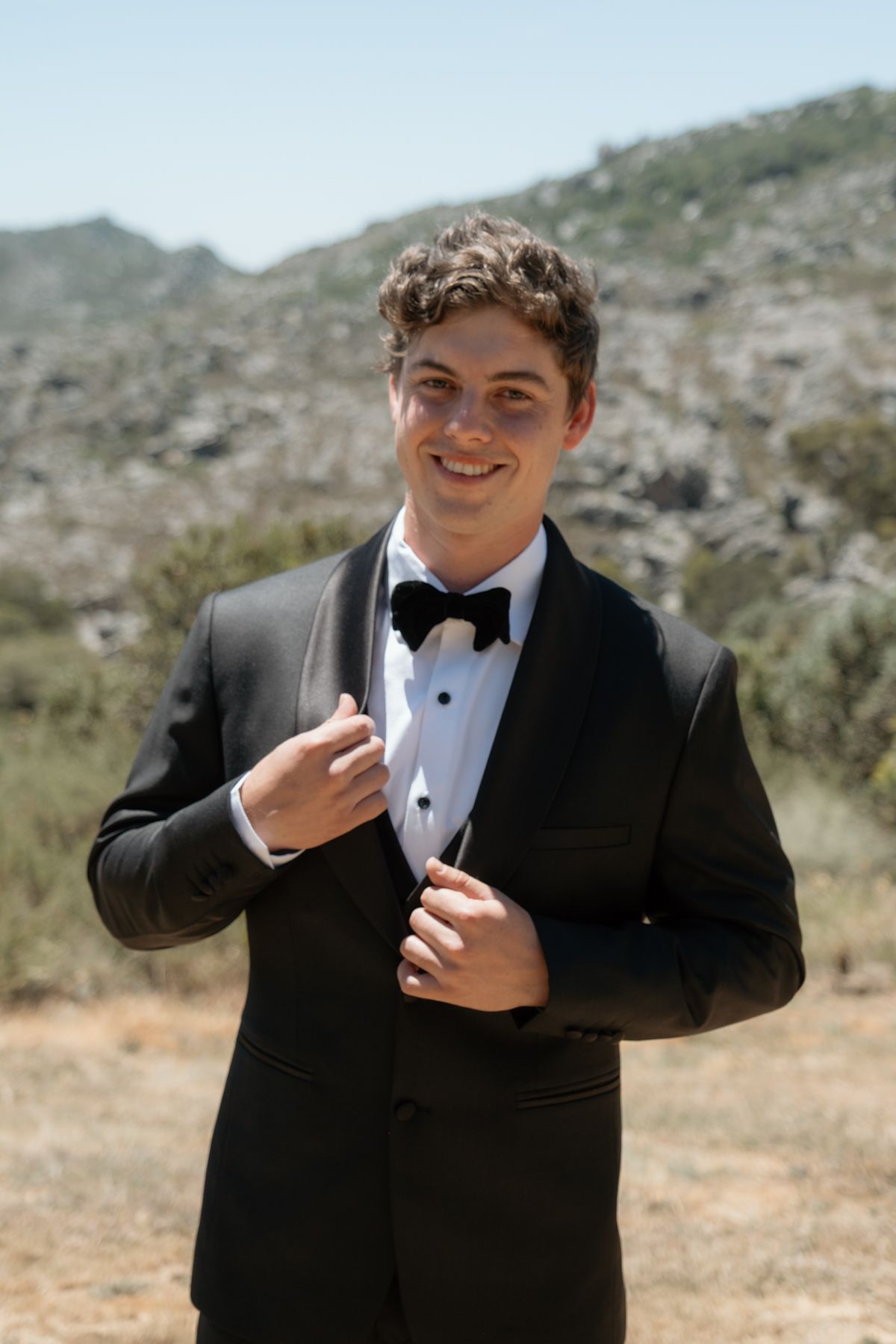 Young groom in black tuxedo with bow tie smiling outdoors with rocky mountain landscape background.