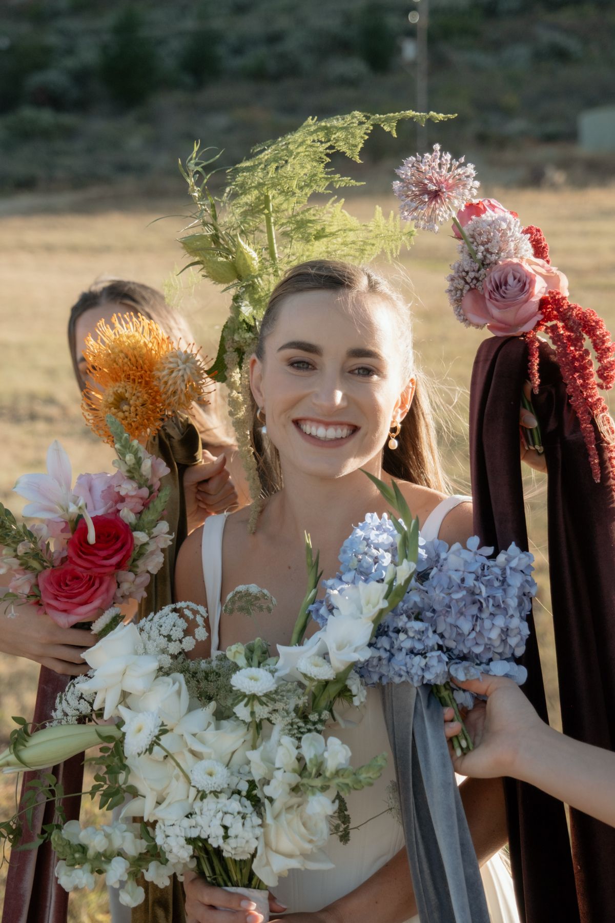 Smiling bride holding a white and blue floral bouquet surrounded by colorful bridesmaid flowers outdoors.