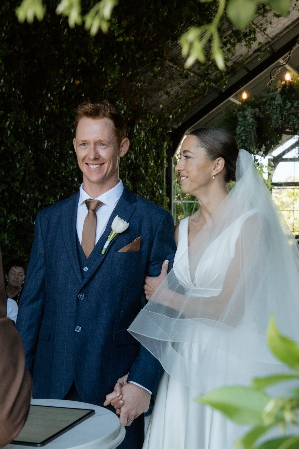 Smiling groom in navy suit holds hands with bride in white veil during outdoor wedding ceremony.