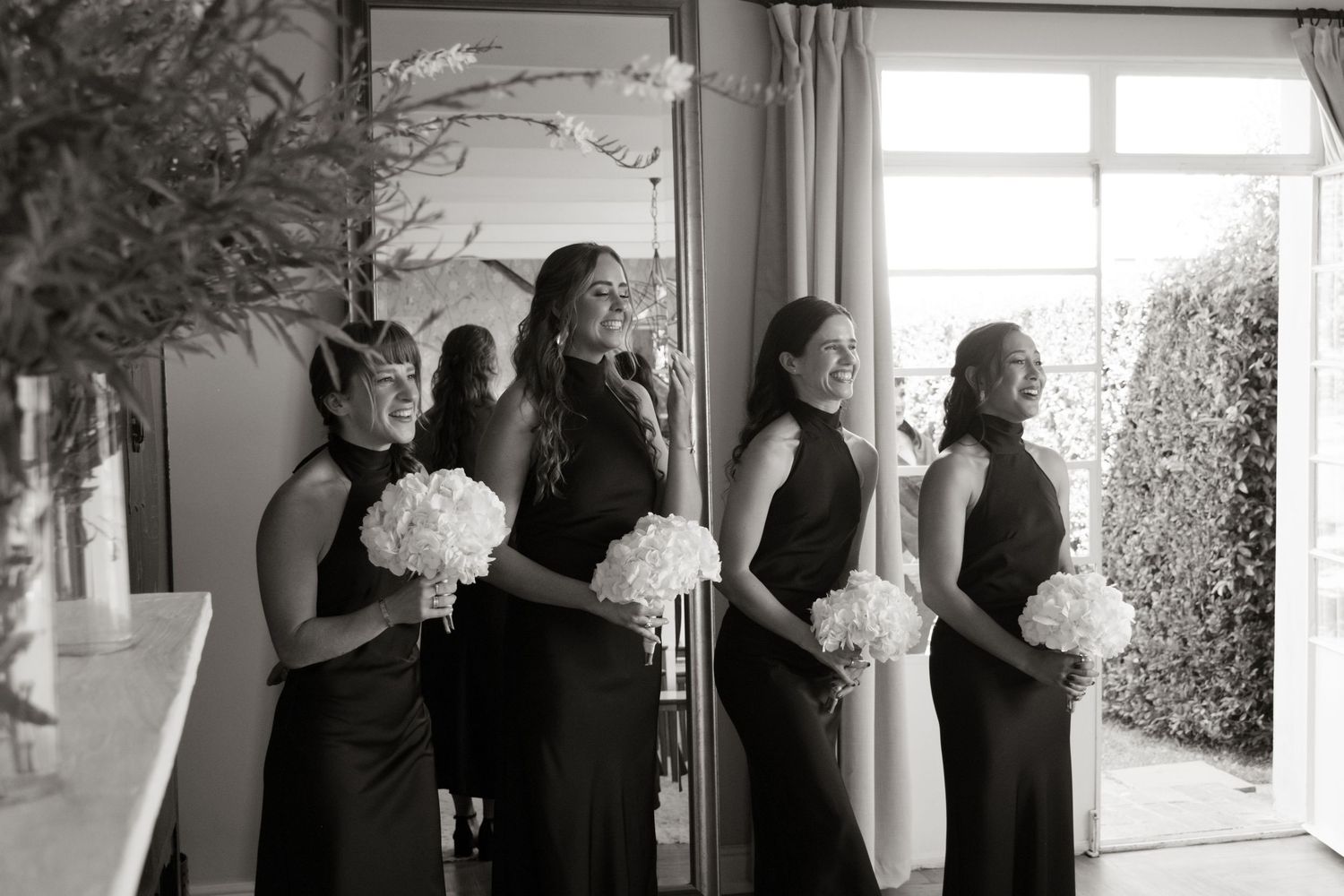 Four bridesmaids in black dresses holding white bouquets laughing together in a bright room before a wedding ceremony.