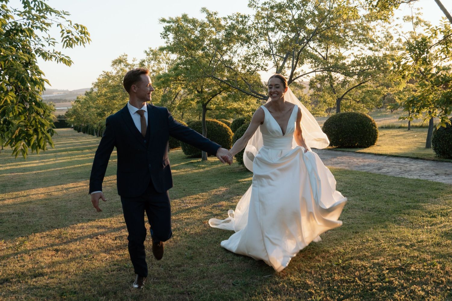 Bride and groom holding hands running through lush green gardens during golden hour at outdoor wedding.