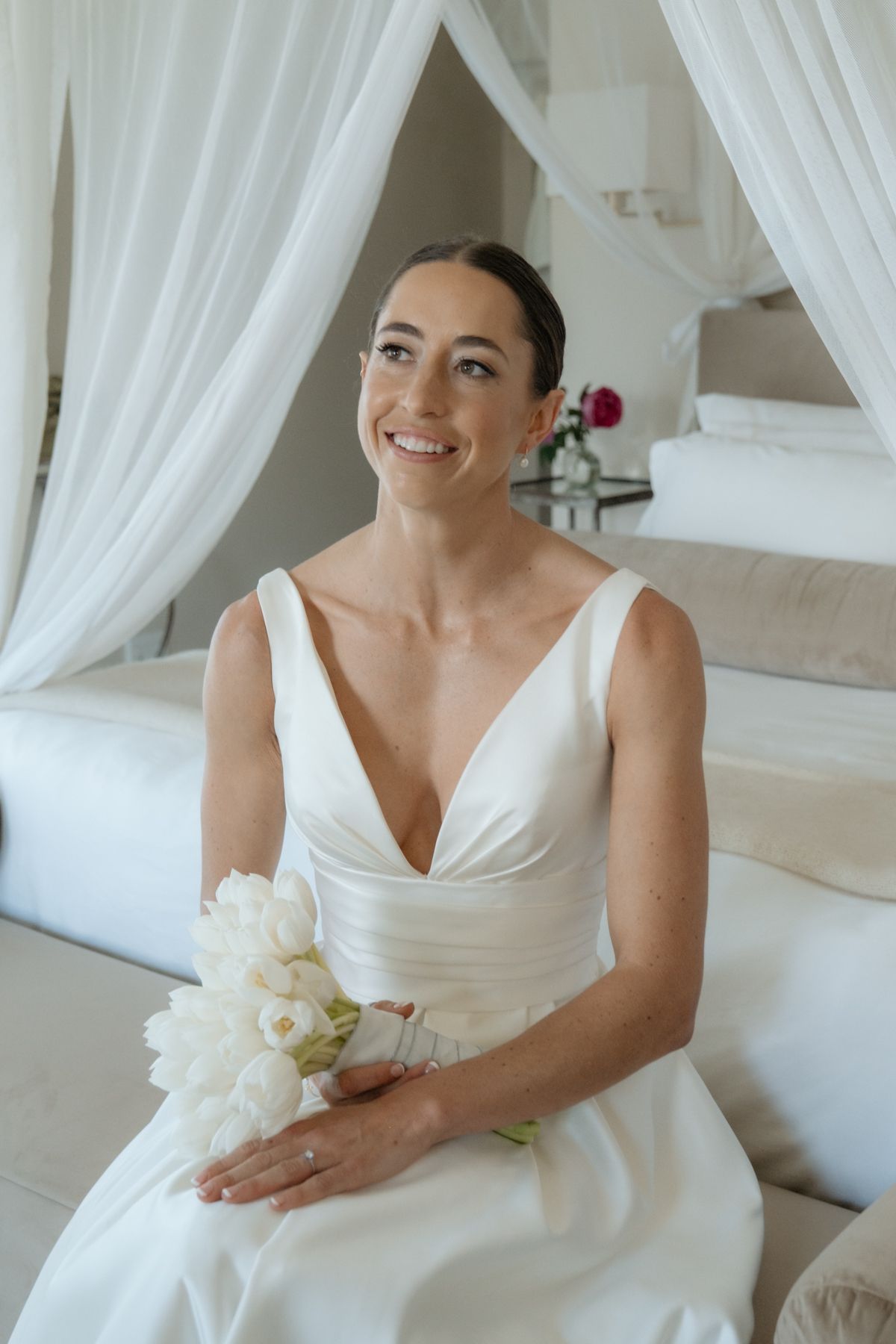 Radiant bride in a white V-neck wedding gown smiling while holding a bouquet of white tulips in a bright room.