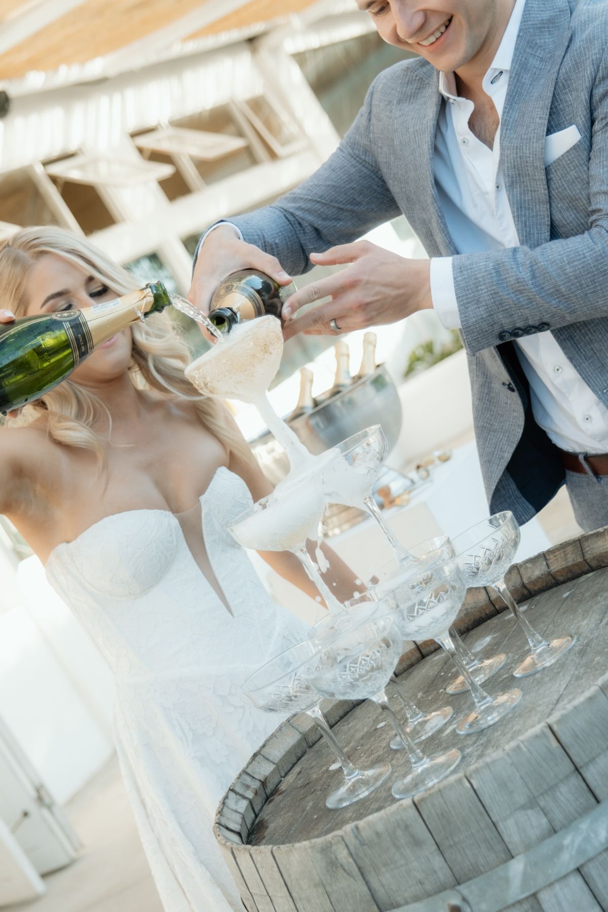 Smiling groom in grey suit pours champagne tower with bride in strapless wedding dress at outdoor reception.
